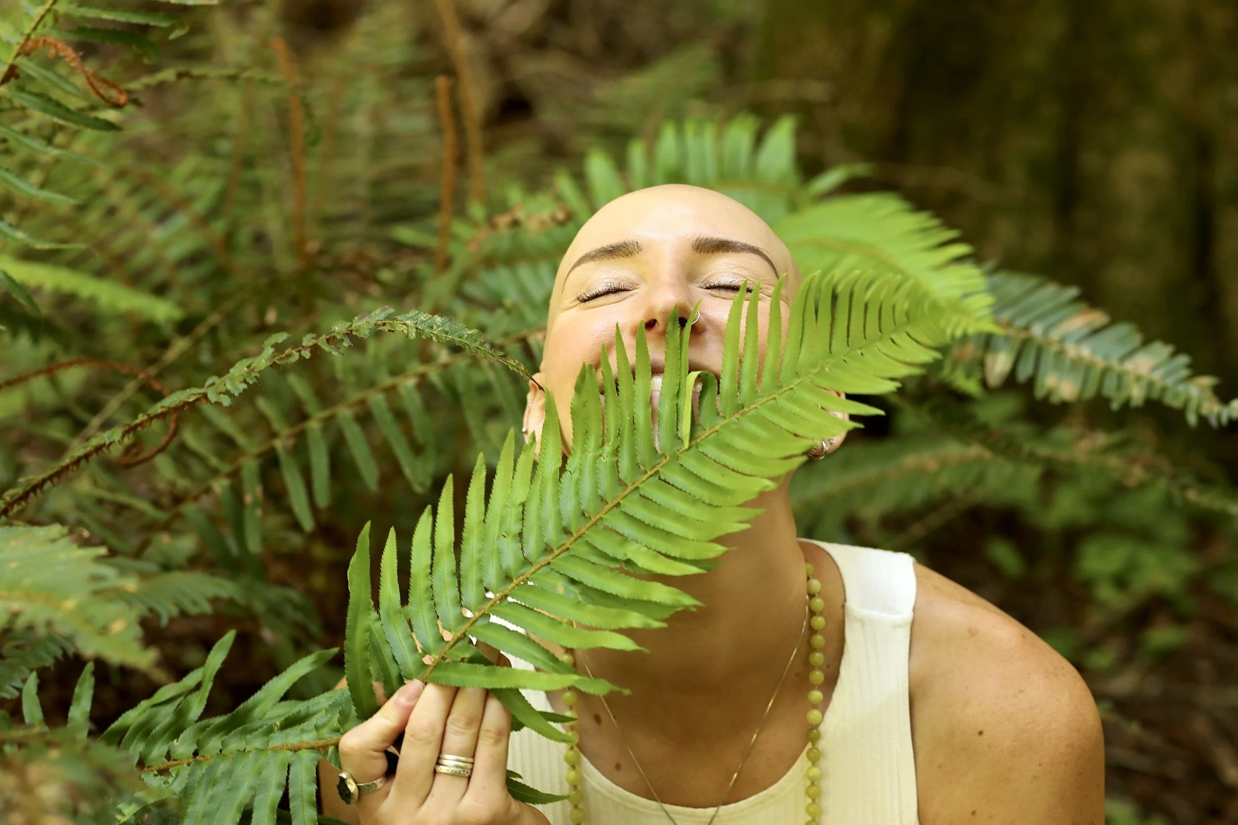 A woman with closed eyes smiling behind green fern leaves in a lush forest.