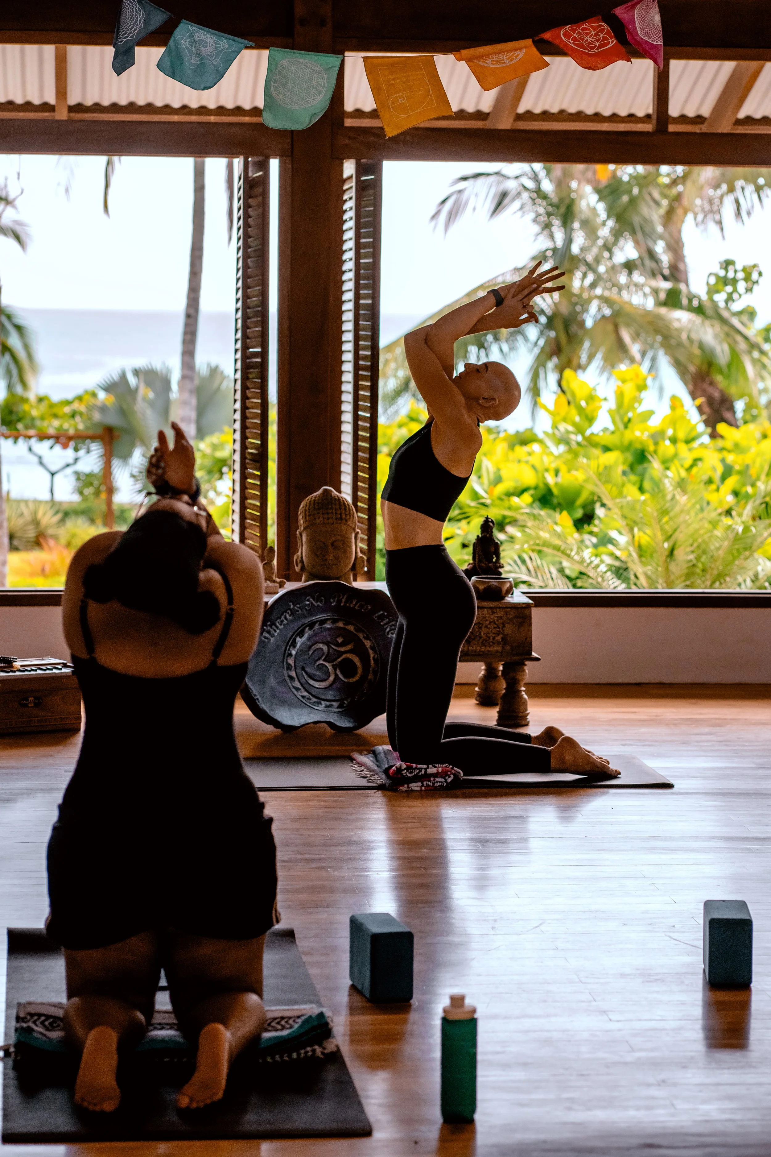 A group of people practicing yoga in a room with large windows opening to a tropical outdoor scenery with palm trees and greenery. One person is in a kneeling yoga pose with hands in a prayer position, facing upward. Others are also in yoga poses, with props like blocks and water bottles visible.