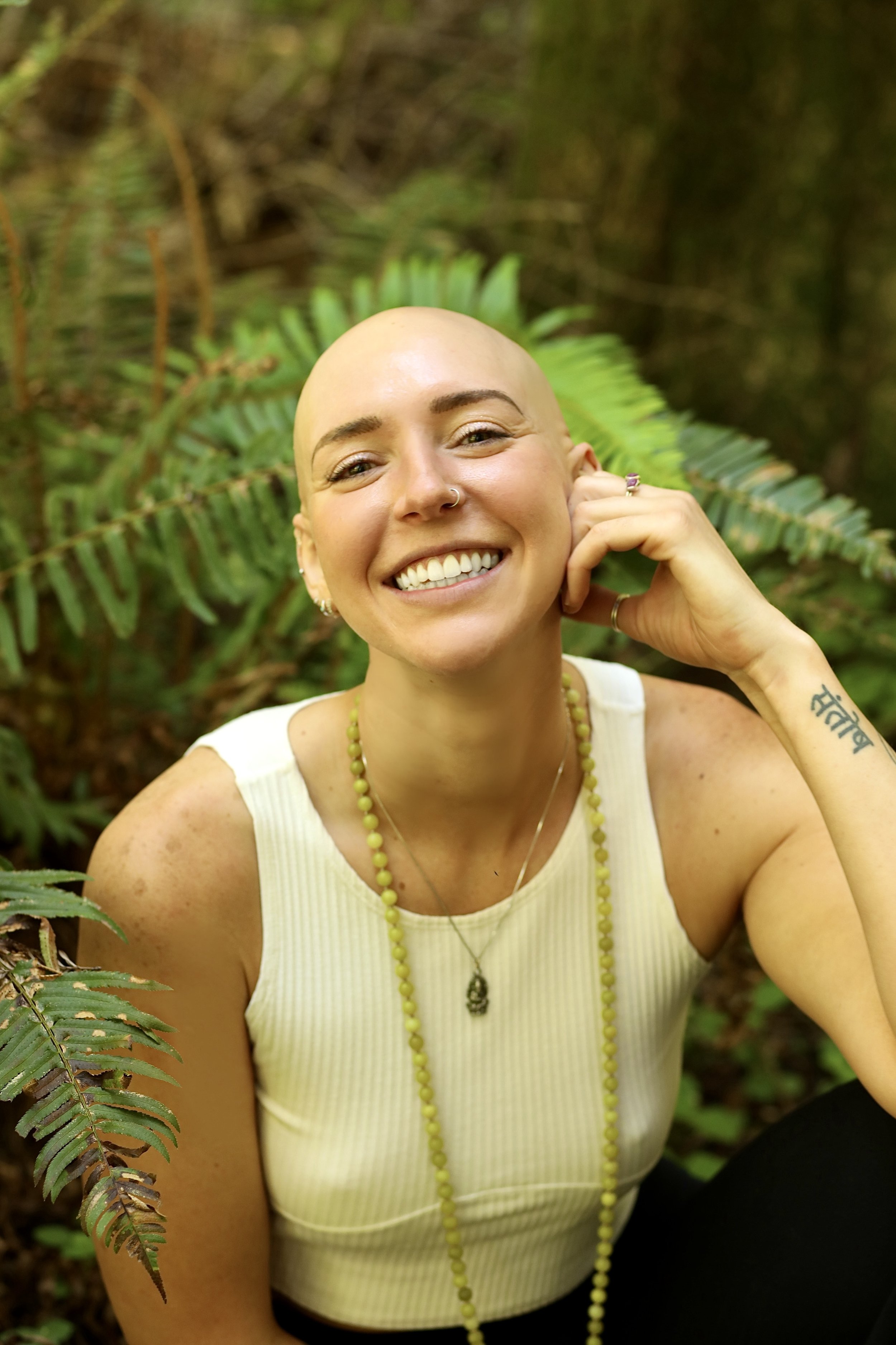 A blonde woman with a shaven head smiling outdoors in front of green ferns, wearing a white sleeveless top, green beaded necklace, and rings.