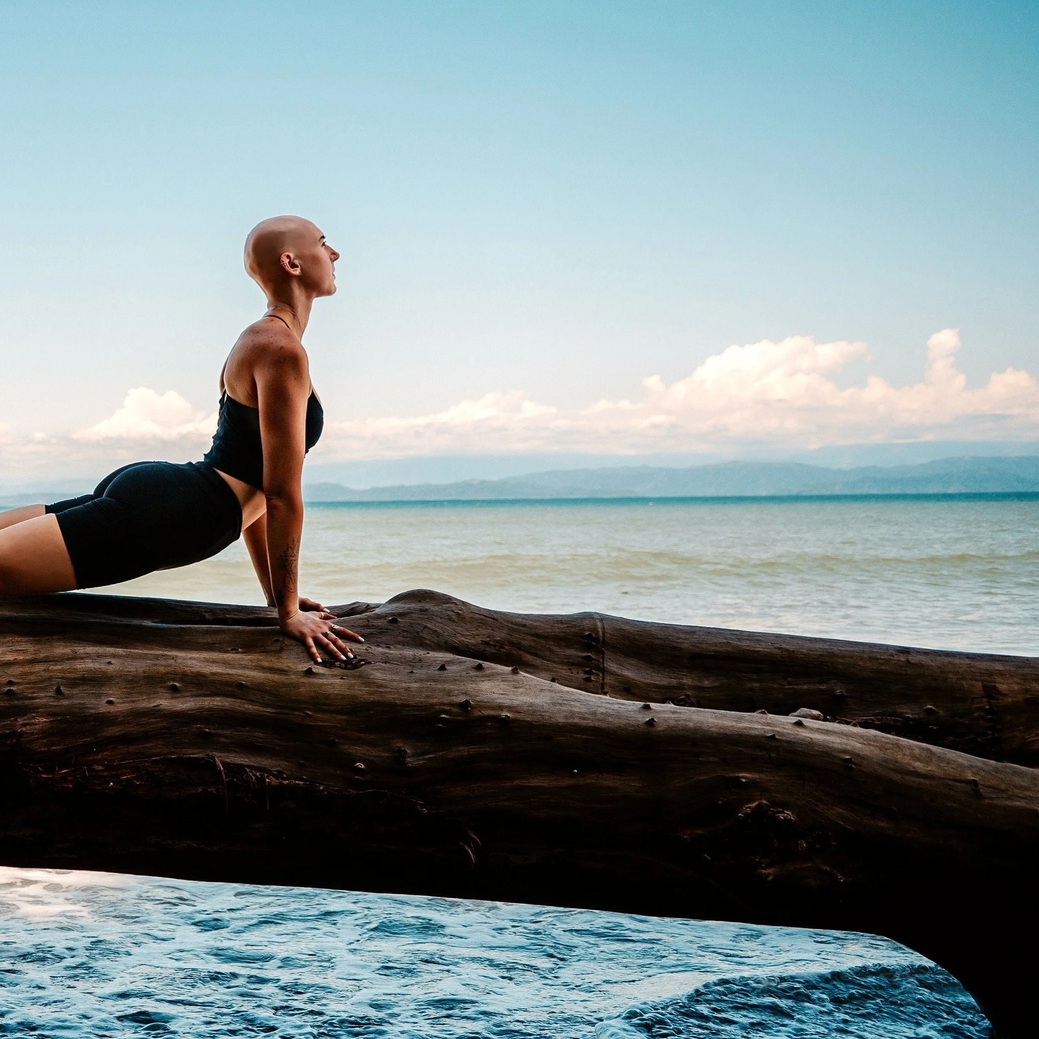 A person practicing yoga or stretching on a large piece of driftwood on a beach, with the ocean and a cloudy sky in the background.