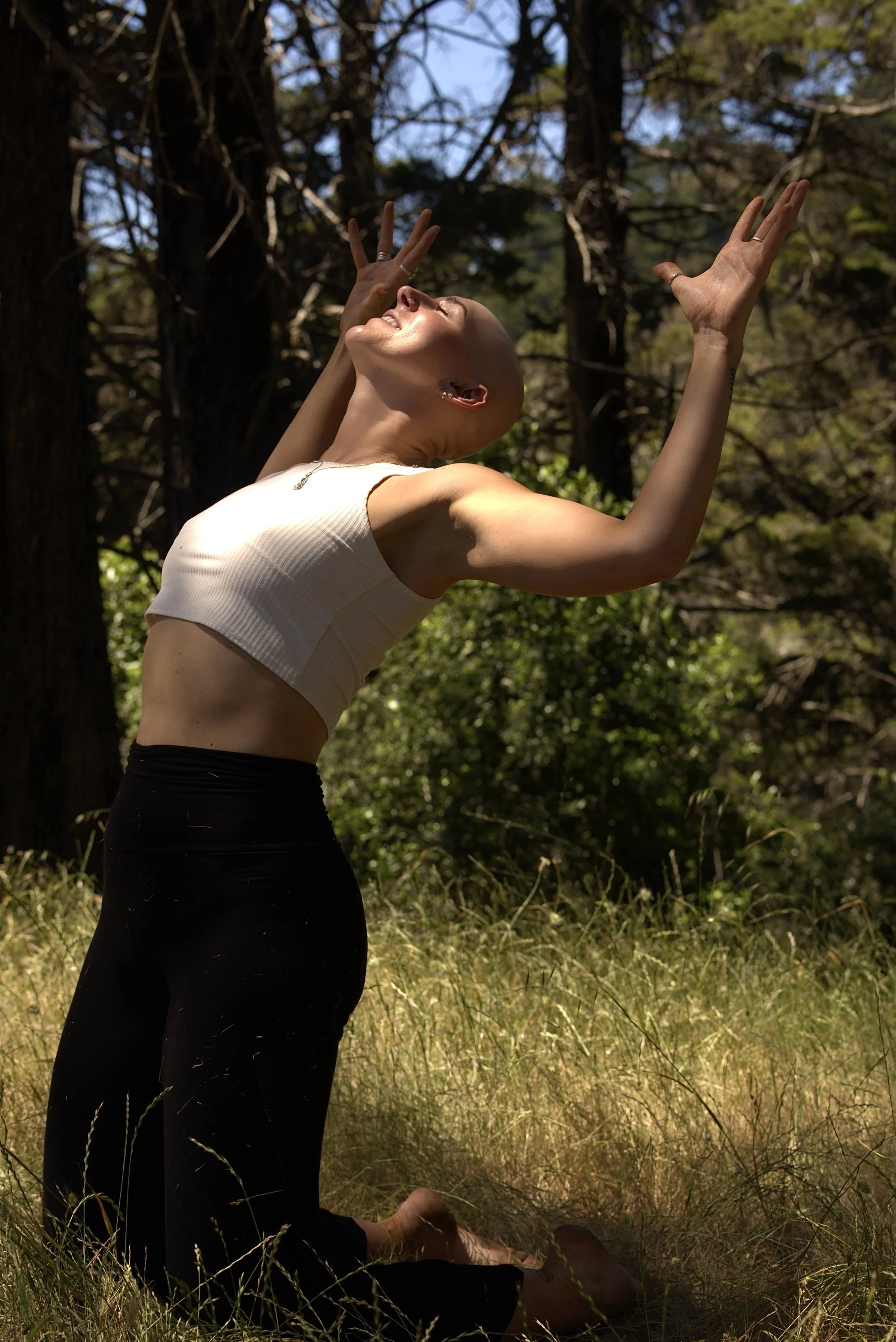 A woman with a shaved head practicing yoga outdoors among trees and grass, sitting in a seated forward bend pose with arms raised and head tilted back.