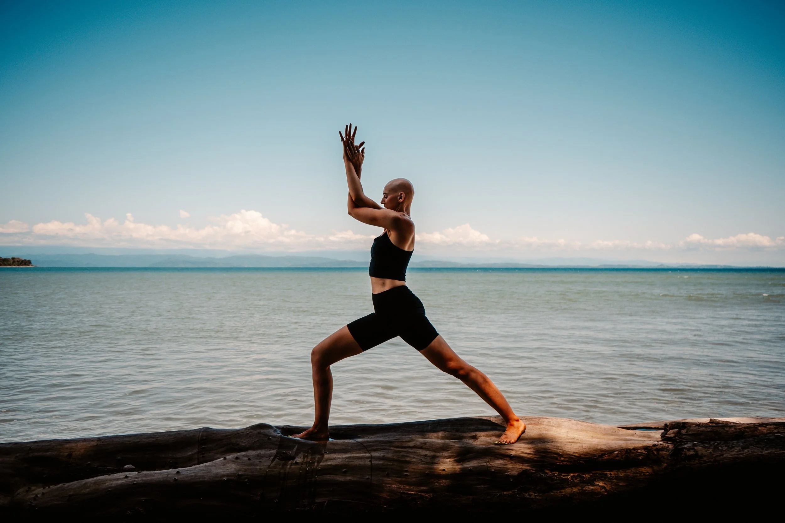 A person practicing yoga outdoors on a log at the beach, standing in a warrior pose with hands in prayer position, with water and mountains in the background.