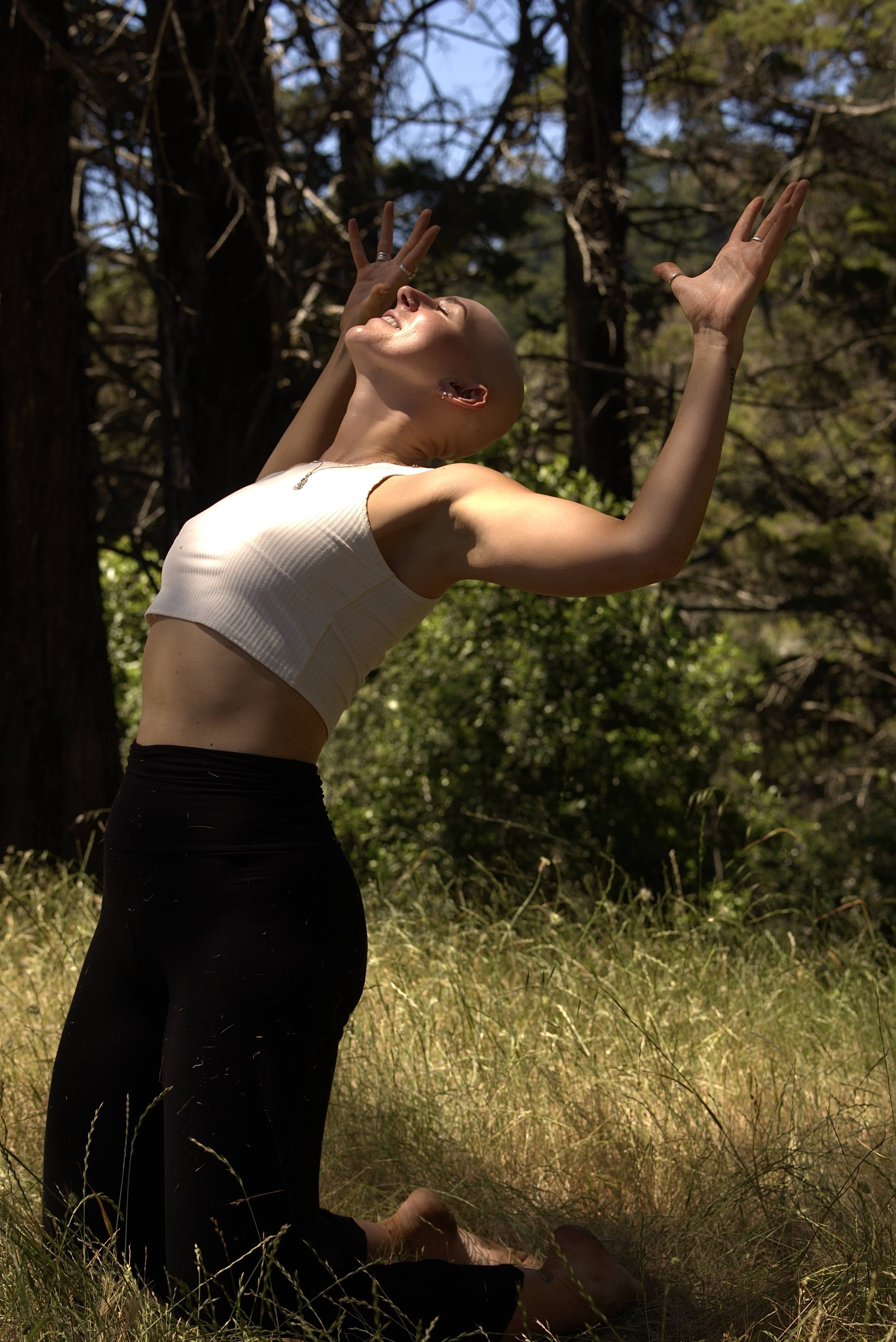 A woman practicing yoga outdoors in a forest, sitting on the grass with her eyes closed, smiling, and performing a pose with her hands raised and fingers spread wide.