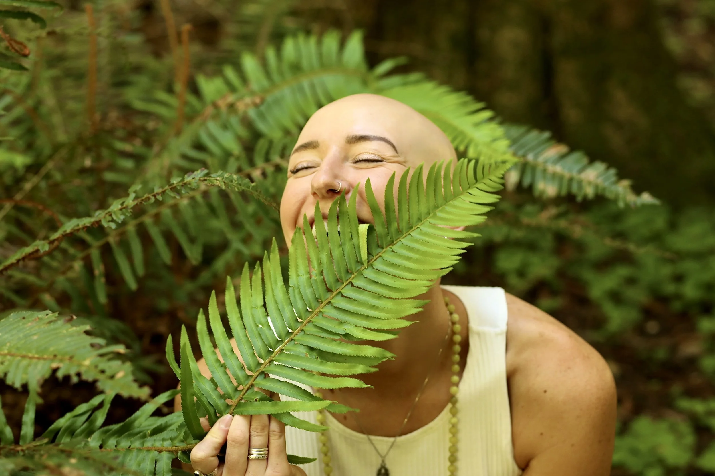 A woman with a bald head, gold makeup, and wearing a yellow dress and necklace is in a forested area, surrounded by green ferns. She is smiling with her eyes closed and holding a fern leaf in front of her face.