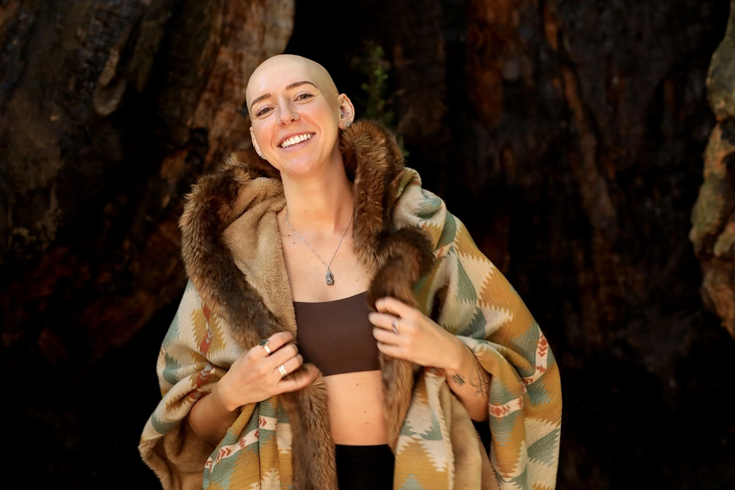 A woman with a shaved head smiling, wearing a silver necklace, a brown top, and a colorful patterned shawl with fur trim, standing in front of a dark, textured background.