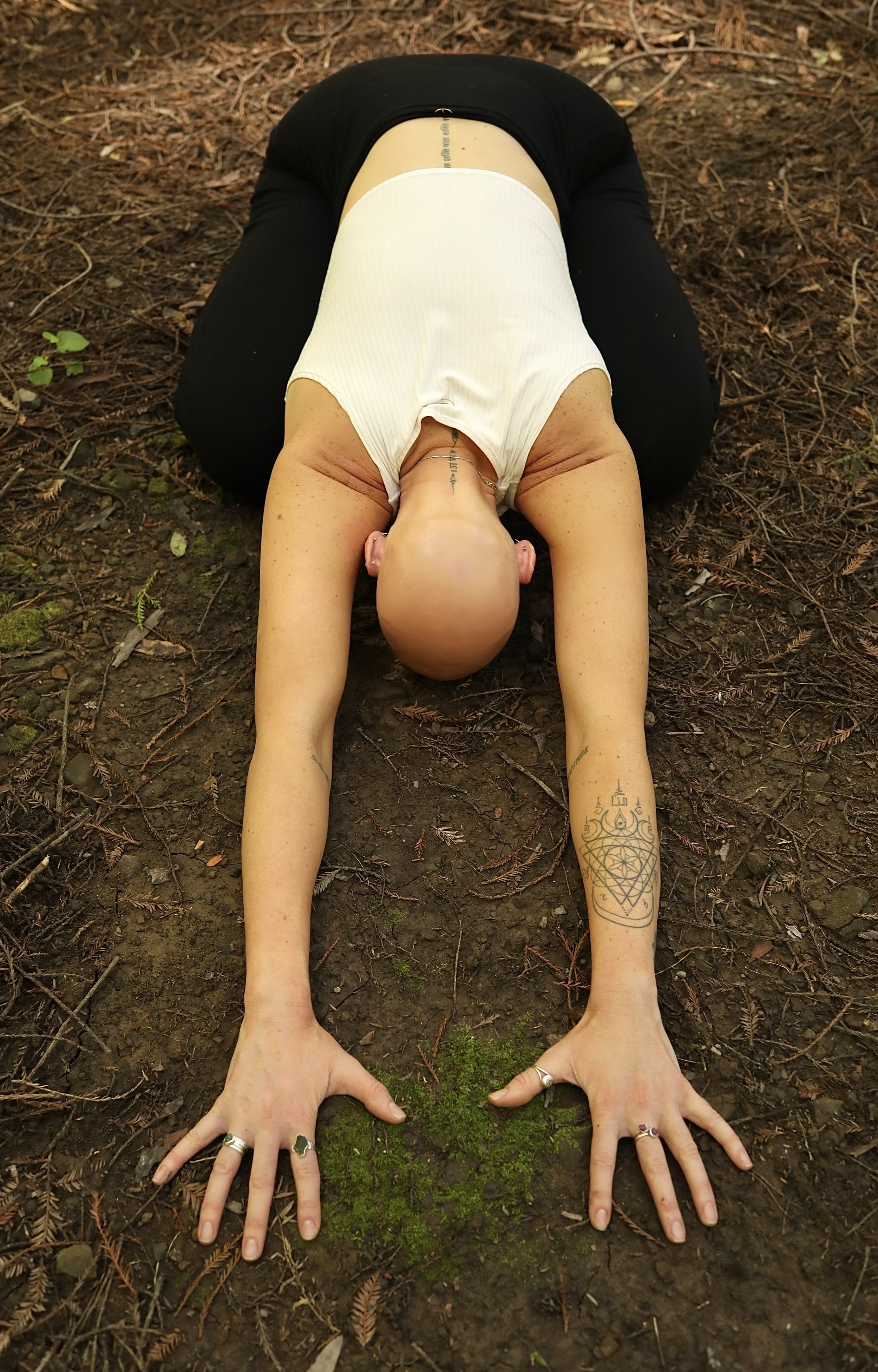 A person's body positioned in a yoga pose on a forest floor, hands on the ground, with tattoos on arms, bald head, wearing a white sleeveless top and black pants.
