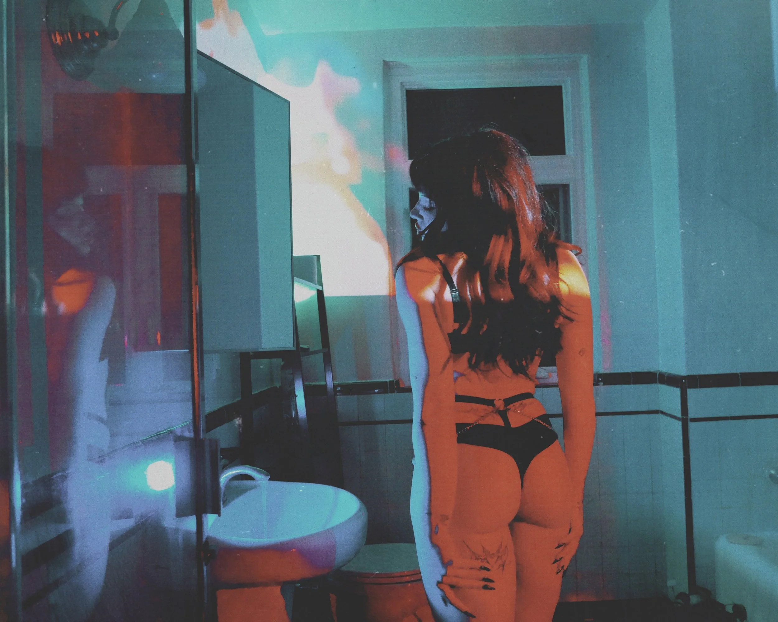 A woman with long hair wearing black lingerie standing in a dimly lit room with colorful lights, looking at herself in a mirror.