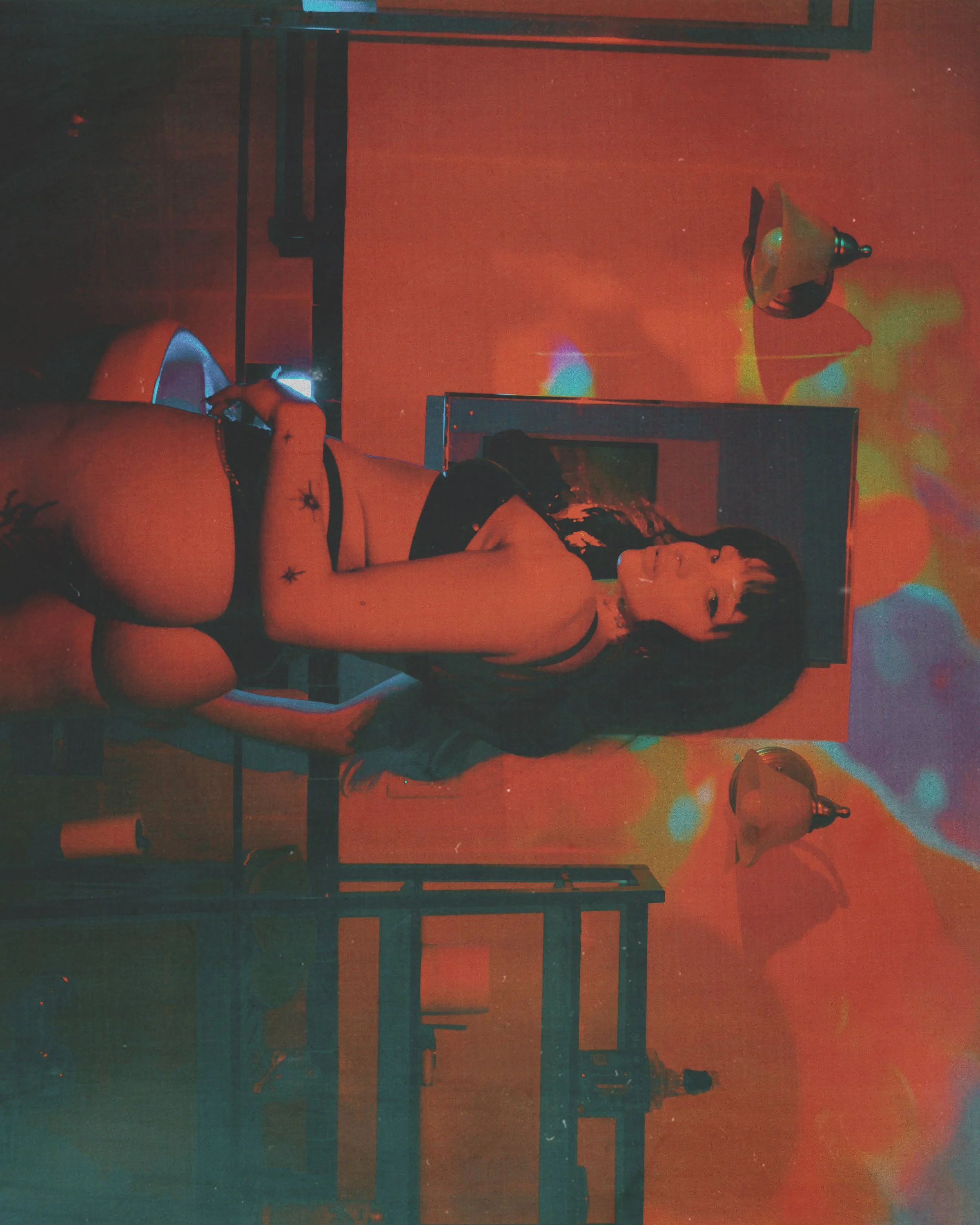 A woman with dark hair and tattoos on her arm, wearing black lingerie, lying on a metal table in a room with red and multicolored lighting, two wall-mounted lamps, a TV, and a picture frame on the wall.
