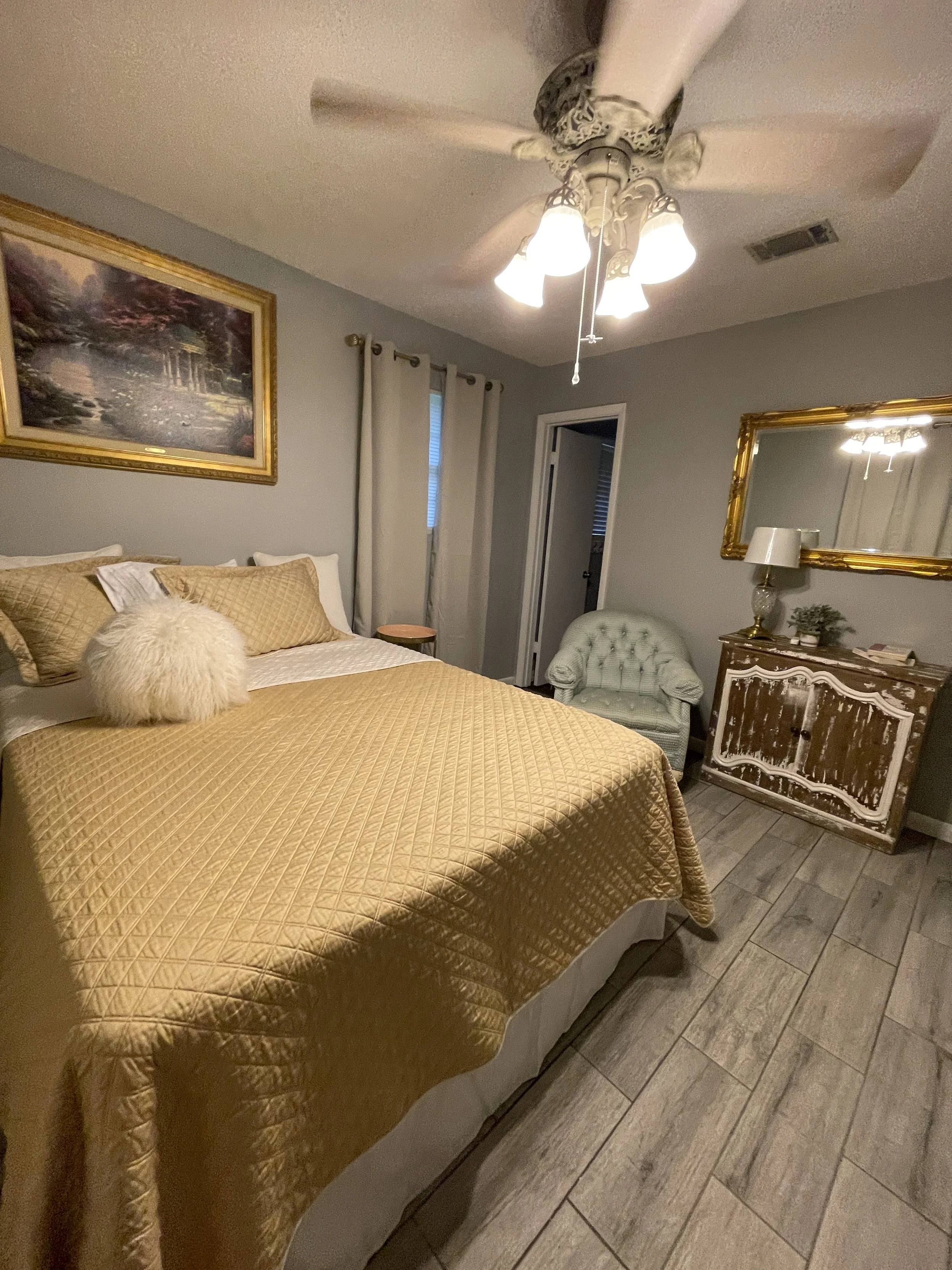 A modern master bedroom featuring a queen-size bed made for two, a ceiling fan, and an accent chair, with a doorway leading to an en-suite master bathroom.