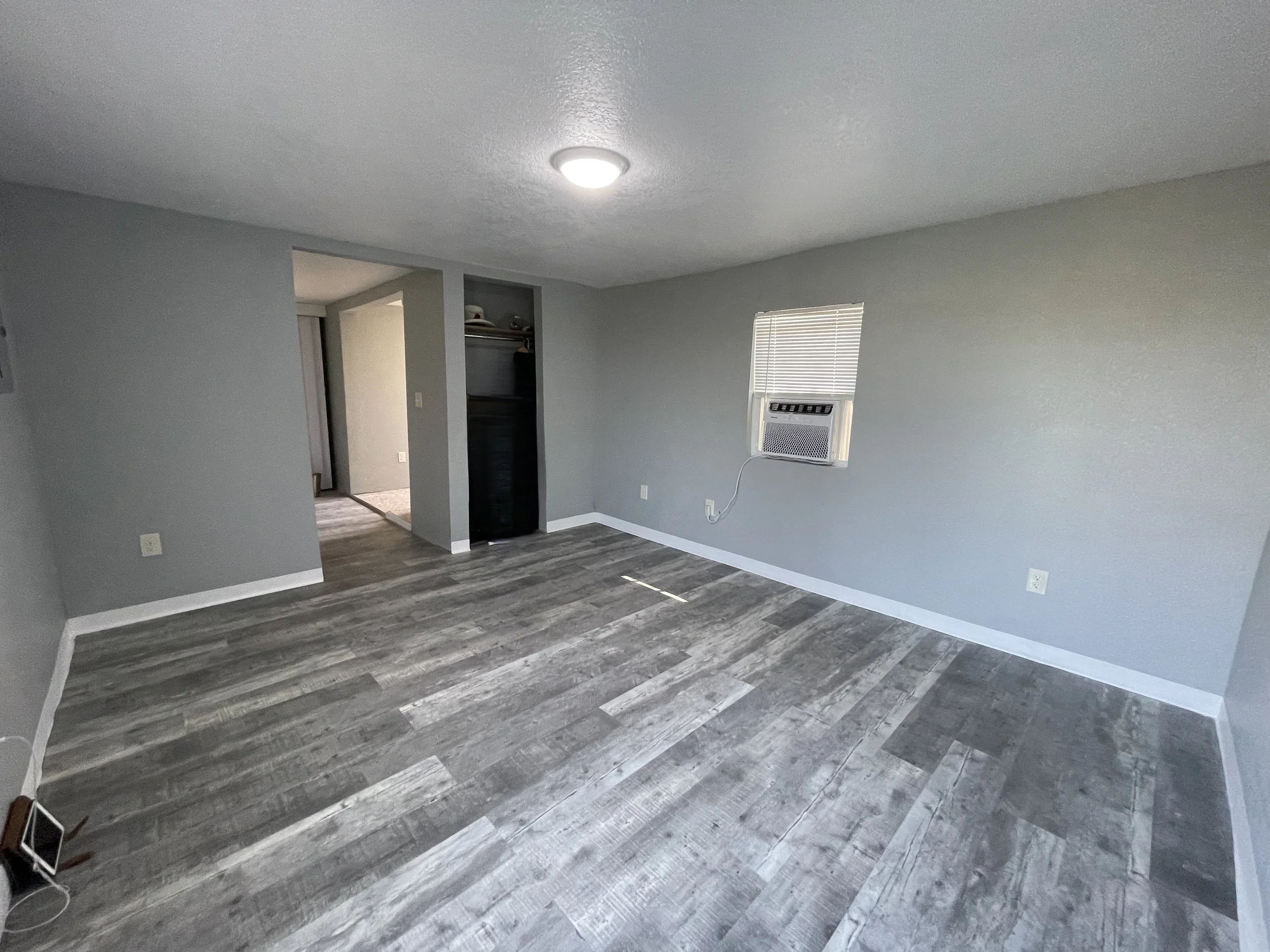 Spacious studio rental room with a dedicated area for a queen bed, armoire, and recliner. Features a through-window heating and cooling unit and an open closet nook.