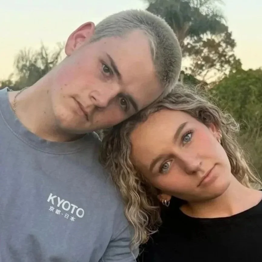 A young man and woman with their heads touching outdoors during sunset, both looking at the camera.