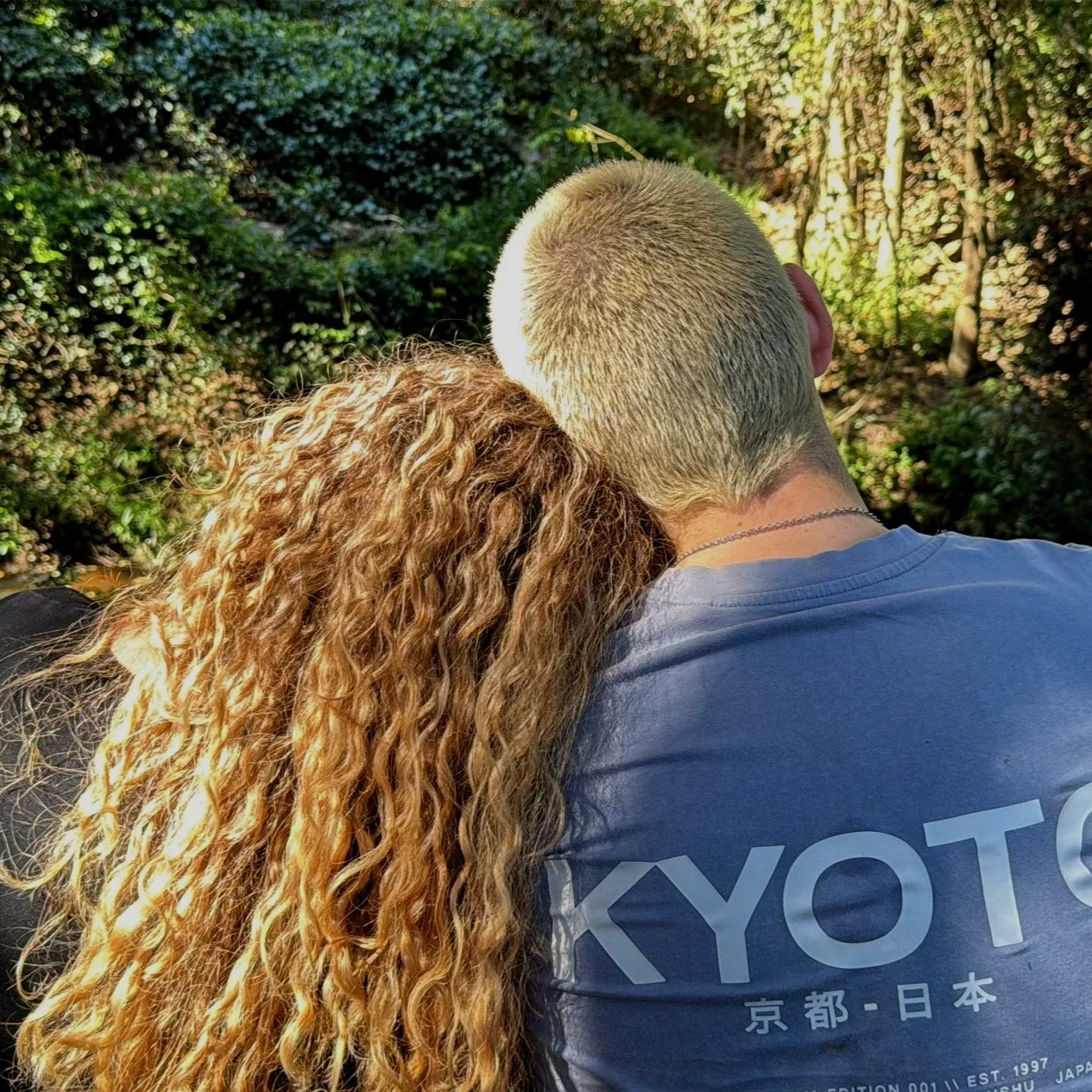 A person with long, curly, blond hair resting their head on the shoulder of a person with a buzz cut, in an outdoor setting with greenery.