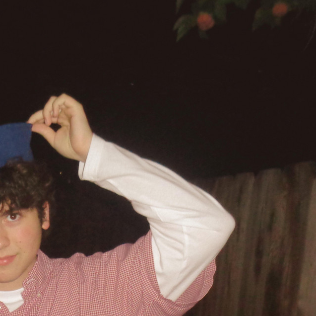 Close-up of a young person making a heart shape with their hand, wearing a red and white checkered shirt and a white long-sleeve shirt underneath, with a dark background and some flowers or plants visible at the top
