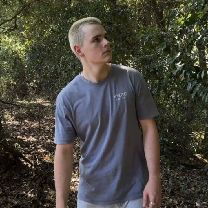 Young man with platinum blonde hair wearing a gray T-shirt with 'Kyoto' printed on it, standing outdoors among trees and bushes.