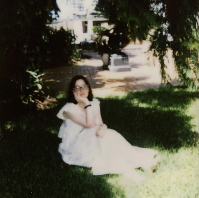 A woman with glasses and dark hair sits on the grass under trees, wearing a light-colored dress, with a hand resting on her chin.