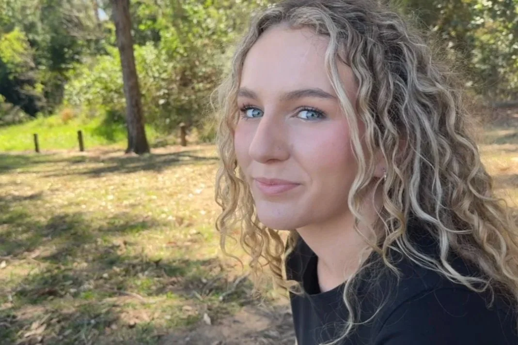 A young woman with curly blonde hair and blue eyes outdoors in a park, looking at the camera.
