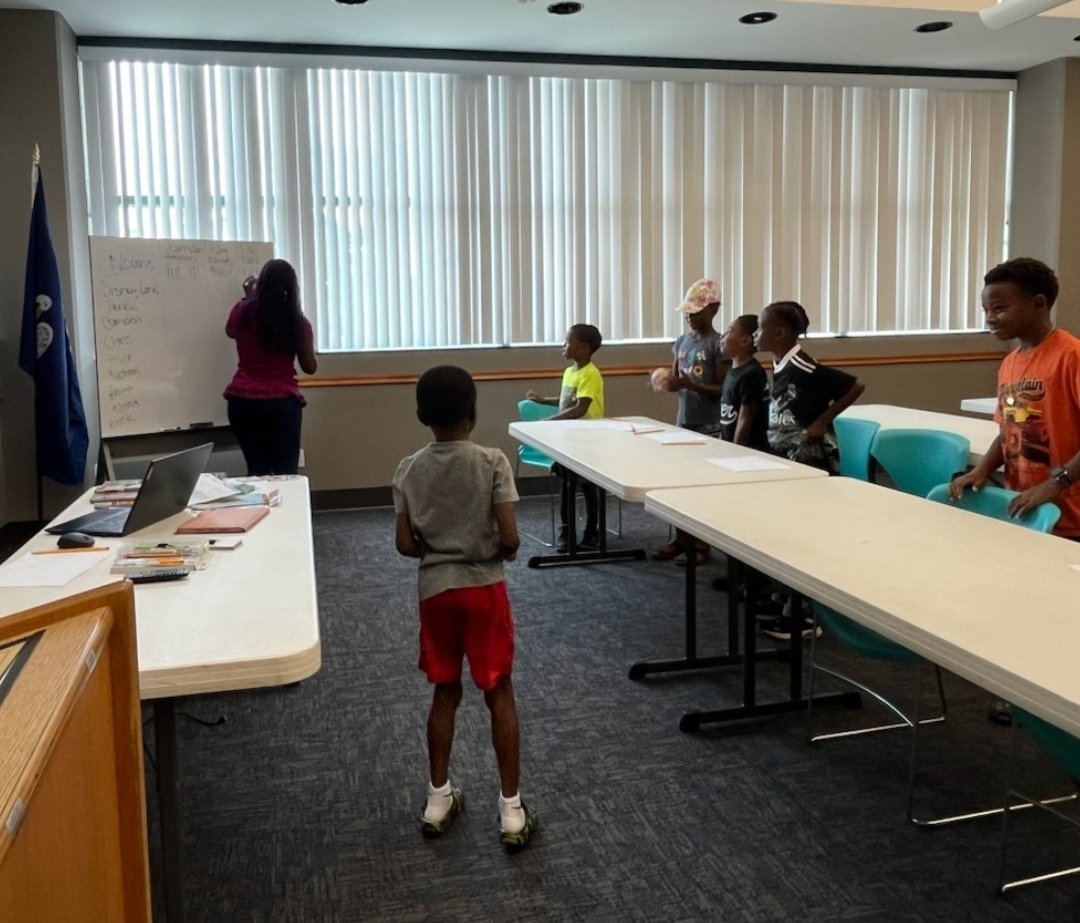 Children participating in a classroom activity, standing in line at the front of the room, while an adult writes on a whiteboard. The room has large windows with vertical blinds, a conference table with laptops and papers, and teal chairs.