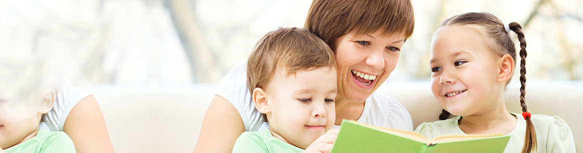 A woman reading a green book with two children, one boy and one girl, all smiling and sitting together on a couch.