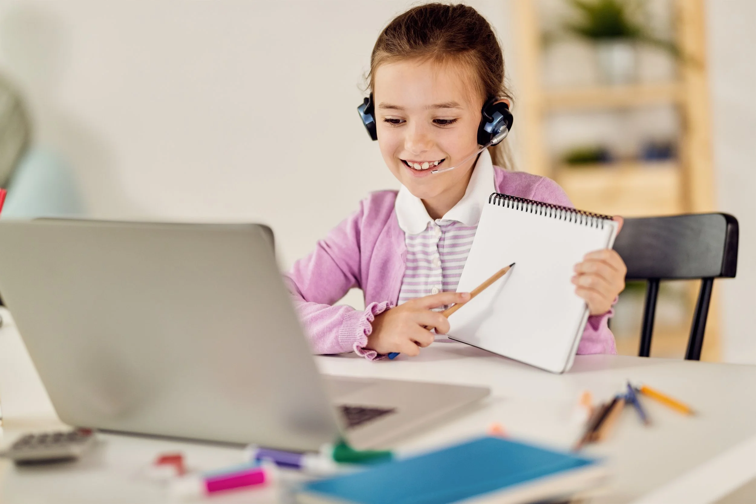 A young girl with a headset and a pink sweater sitting at a desk with a laptop, holding a notepad and smiling.