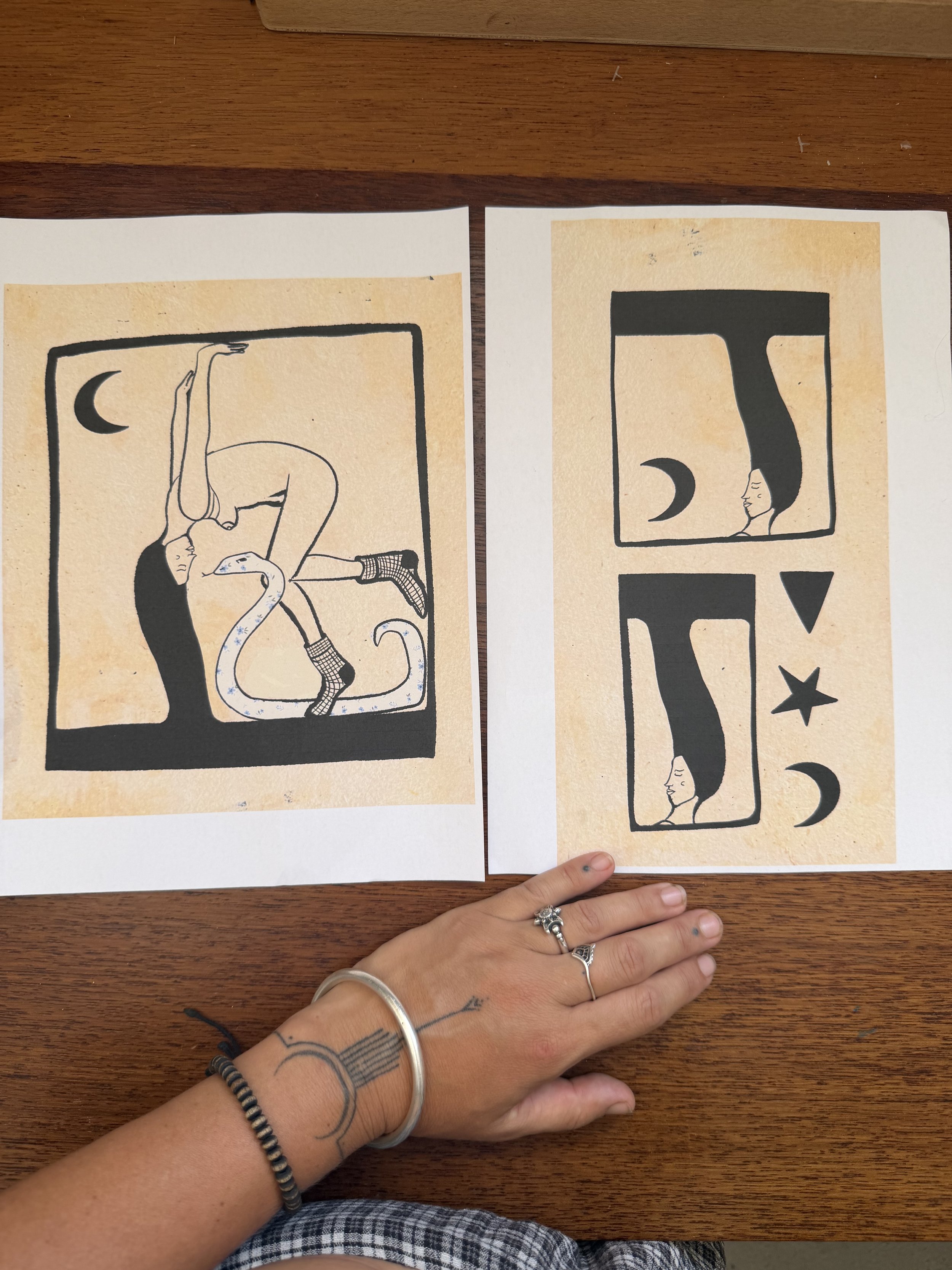 Two pieces of artwork on paper placed on a wooden surface. The left artwork depicts a woman with dark hair, in a curled pose, with abstract elements, a crescent moon, and a snake. The right artwork shows a woman's face with long hair in profile, paired with three zodiac symbols: Aries, Taurus, and Gemini.