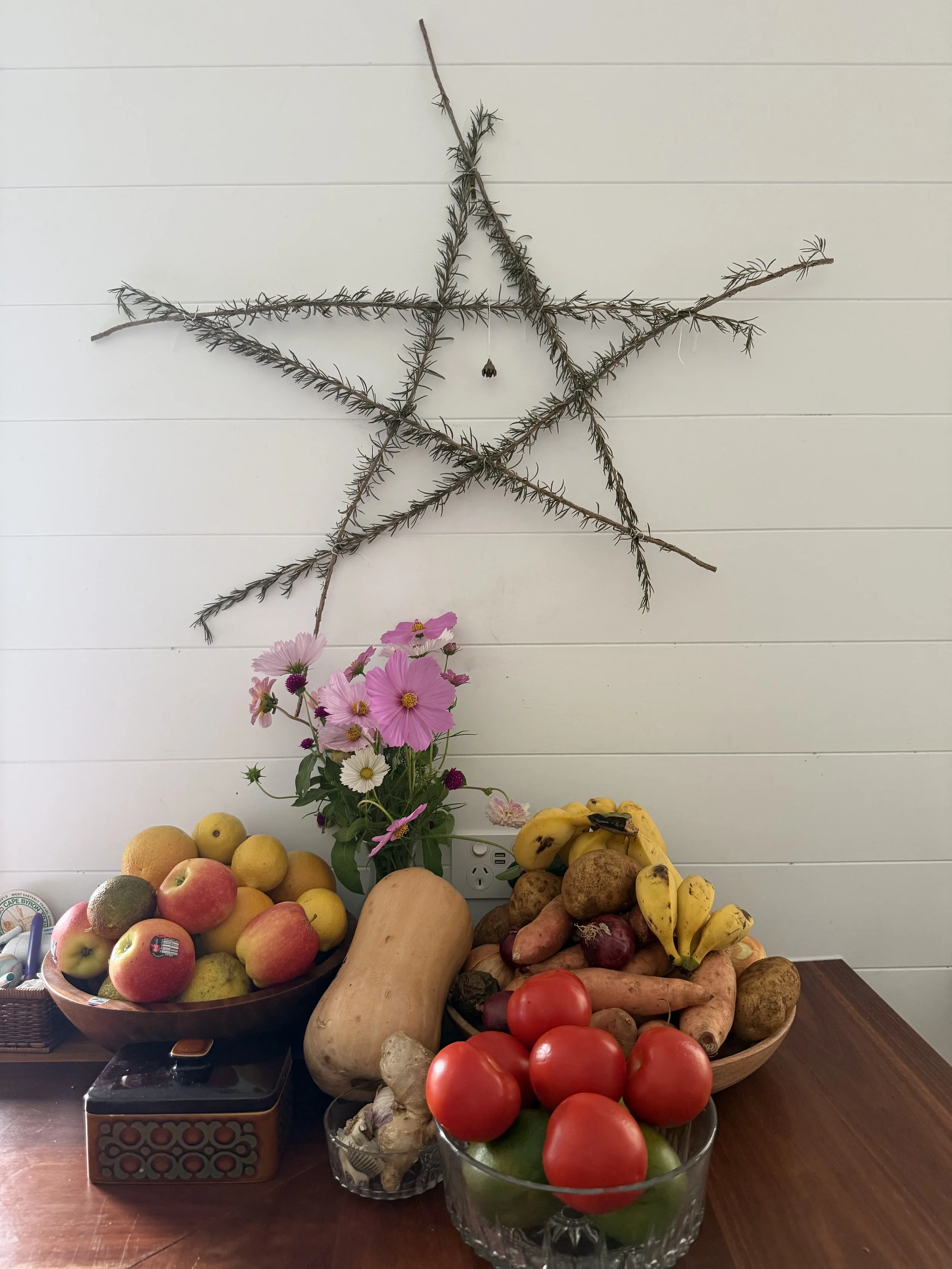 A variety of fresh fruits and vegetables arranged on a wooden table with a vase of pink and white flowers, a star-shaped decoration made of pine branches on a white wall in the background.