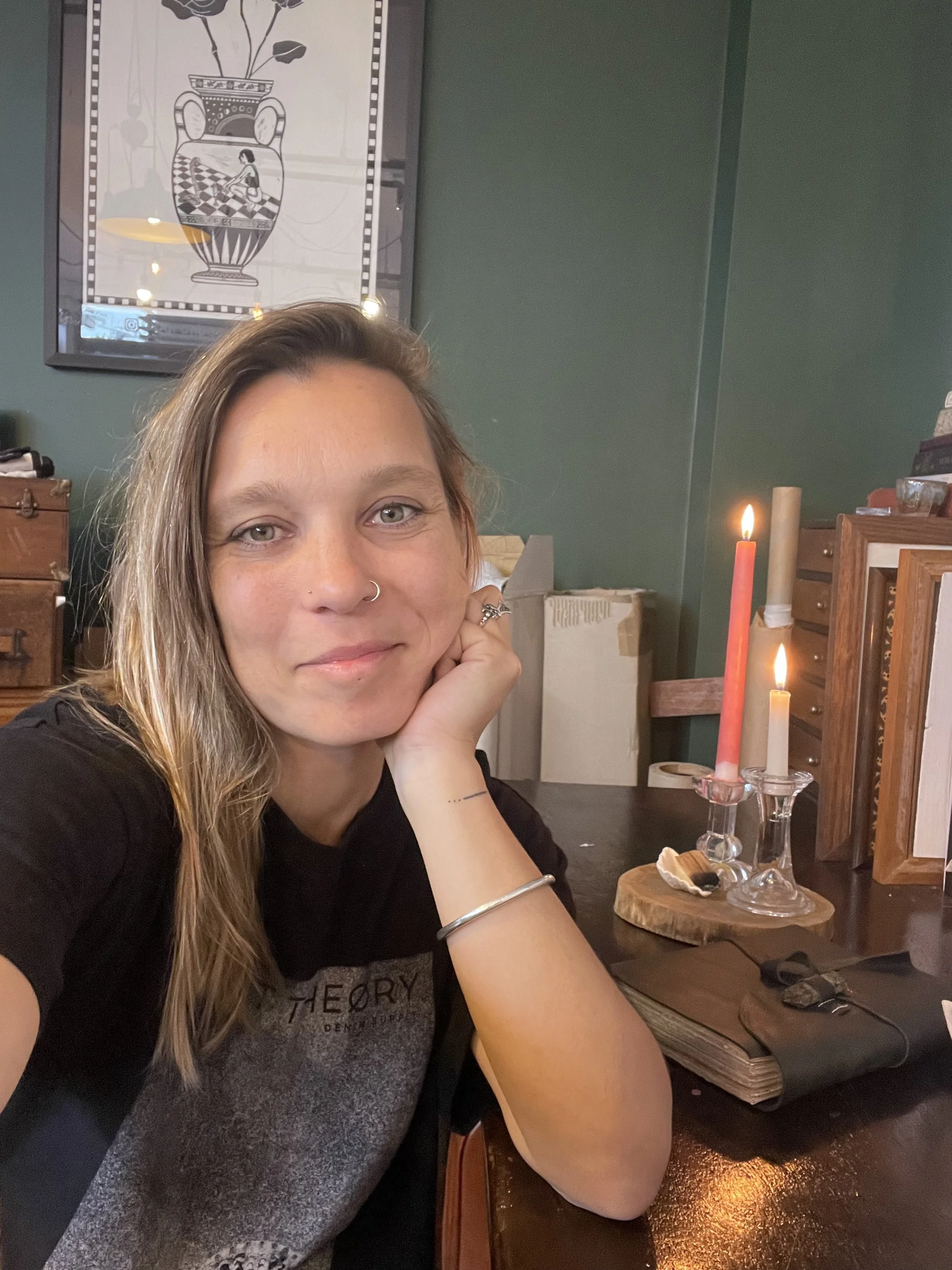 A woman with long hair, wearing a black shirt and jewelry, smiling at the camera, sitting at a desk with lit candles and office supplies in a cozy, warmly lit room.