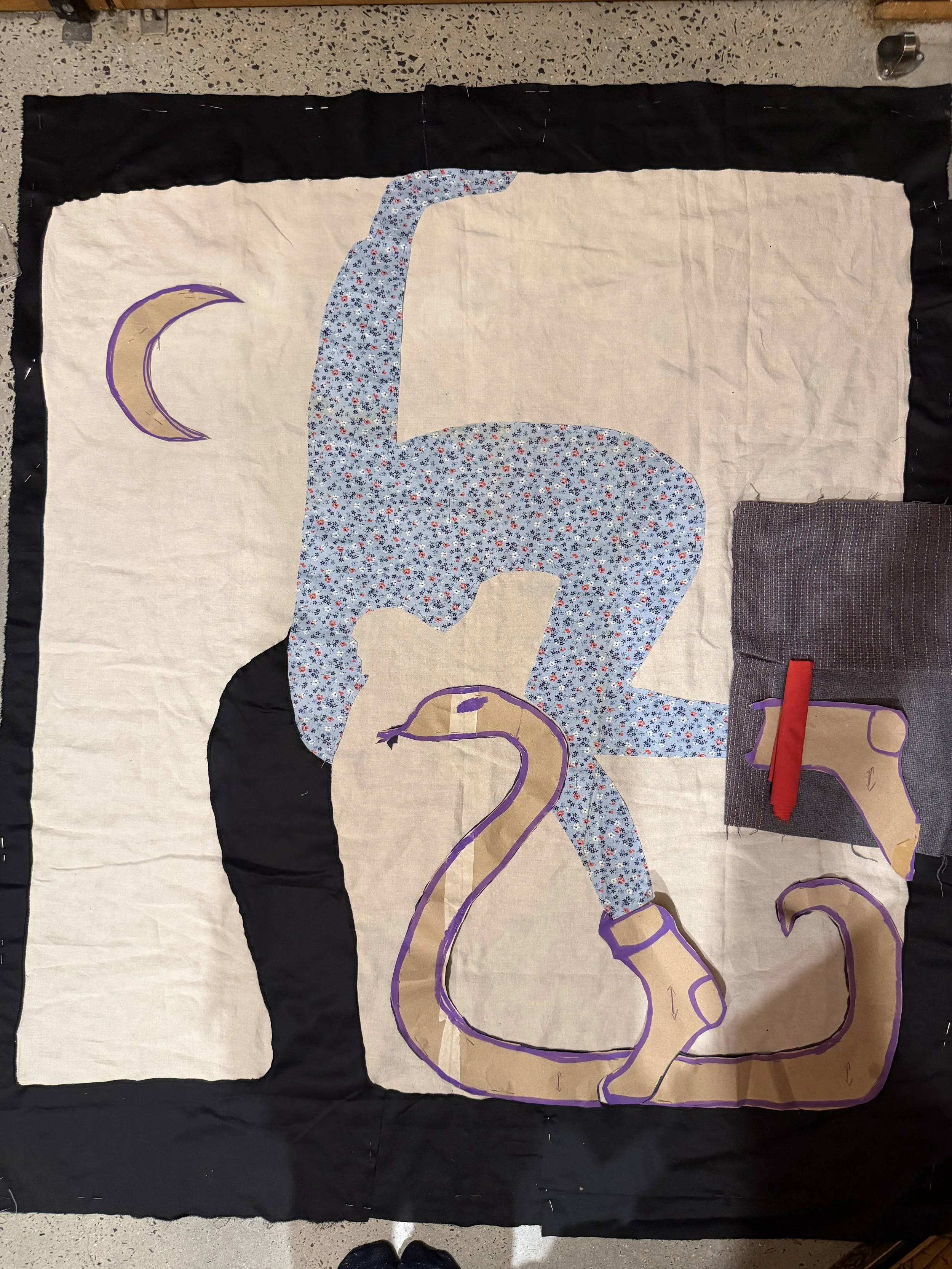 A fabric quilt in process featuring a design of a painter wearing blue floral pants and a black shirt, with a snake outline on a smaller piece of fabric, all laid out on a surface with sewing materials around.