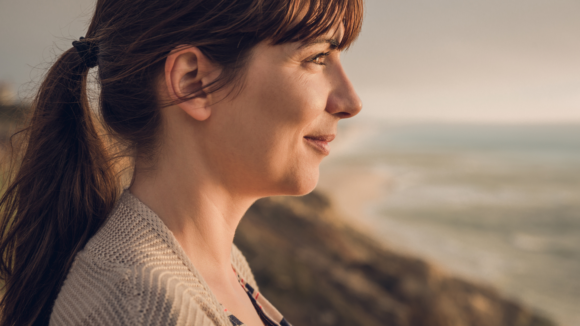 A woman with long brown hair tied in a ponytail looking at the ocean during sunset.