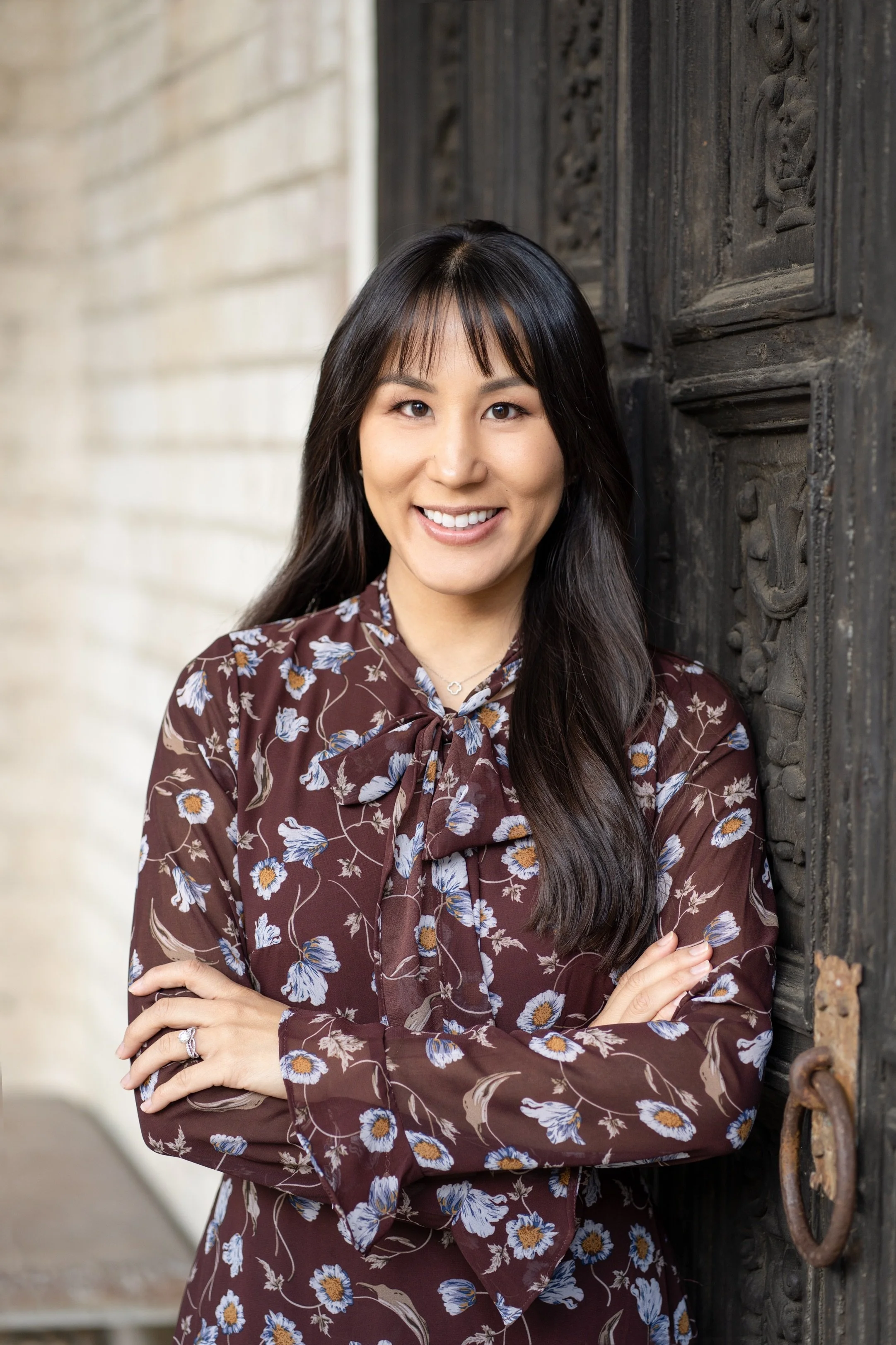 A woman with long dark hair, wearing a brown floral blouse with a bow tie, smiling and standing beside a dark carved wooden door.