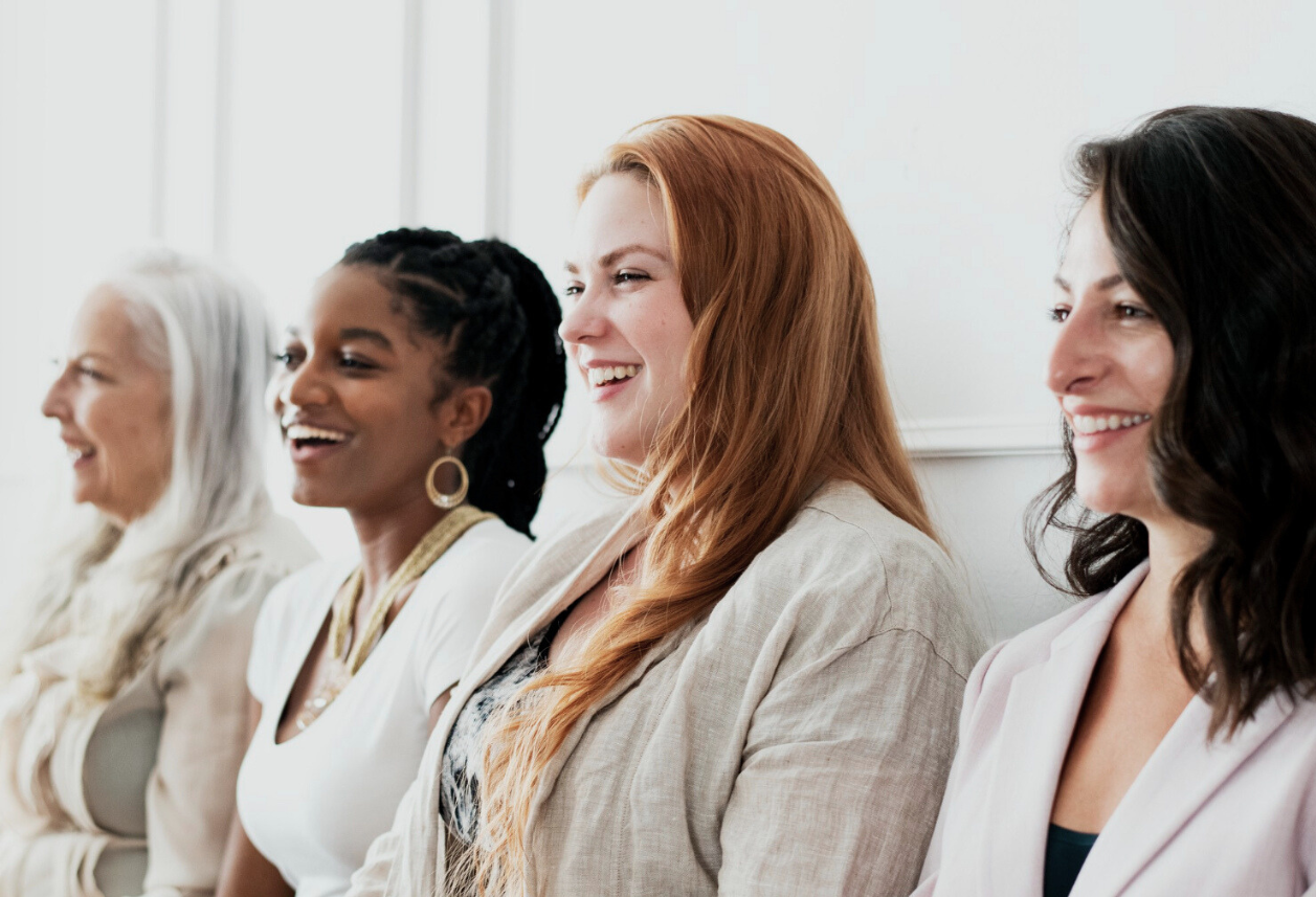 Four women smiling and sitting next to each other at an indoor event.