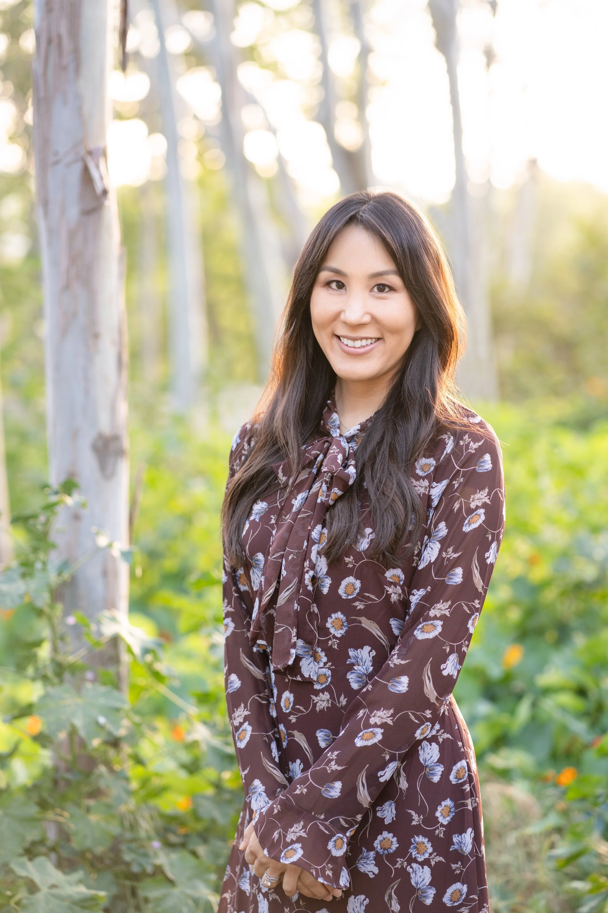 A woman with long dark hair, wearing a brown floral dress, smiling outdoors in a forest with trees and greenery in the background.