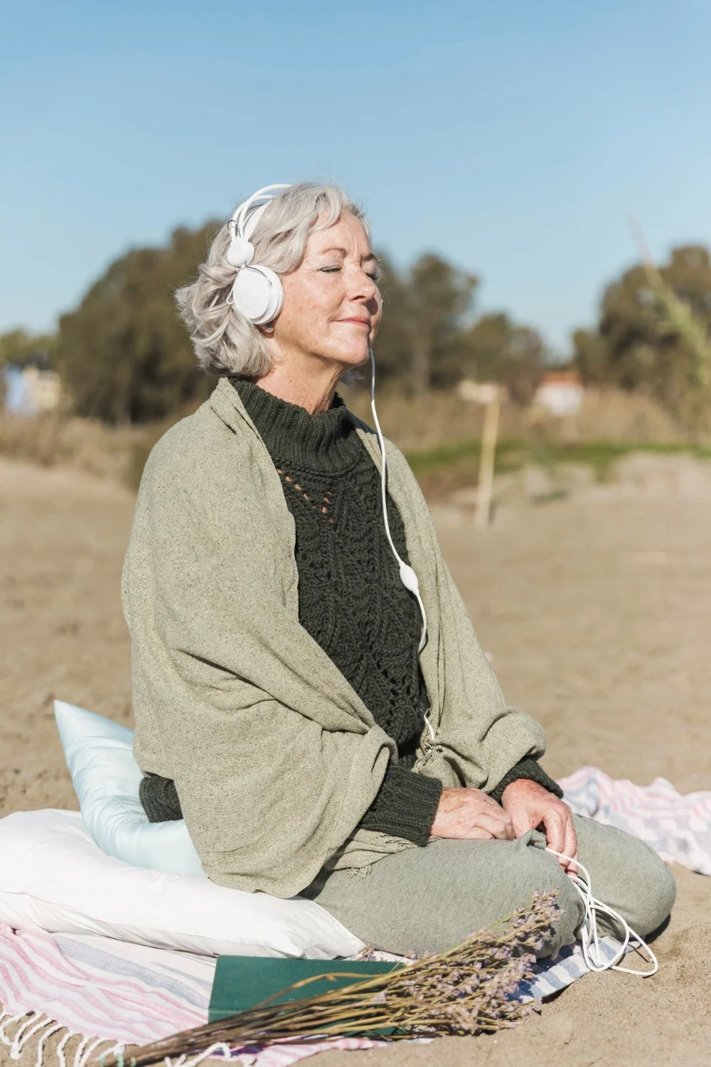 An elderly woman with gray hair, closed eyes, and a peaceful expression, sitting outdoors on a blanket with her eyes closed, listening to music through over-ear headphones. She is dressed warmly in a dark sweater, light jacket, and gray pants, with a pillow supporting her back. The background features a sandy area with trees and a clear blue sky.
