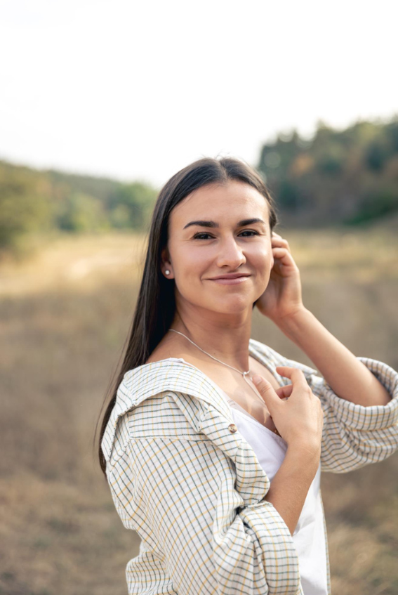A young woman with long dark hair smiling outdoors in a natural setting, wearing a checkered shirt and a white top.