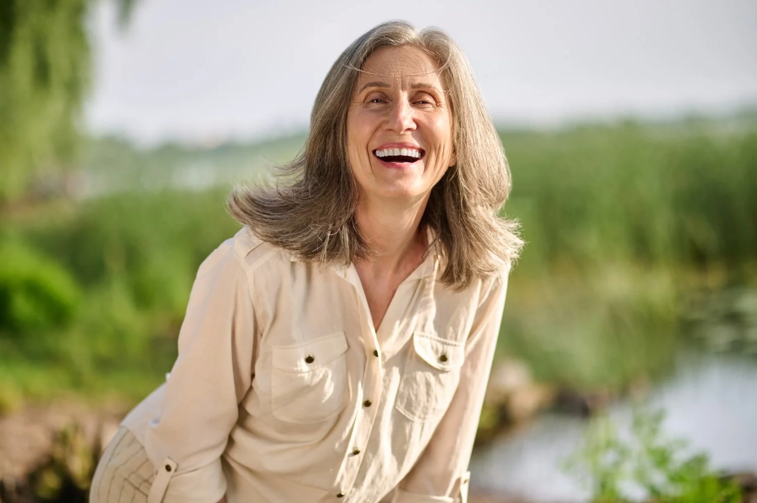 Smiling elderly woman with gray hair in an outdoor natural setting near water, wearing a beige shirt.