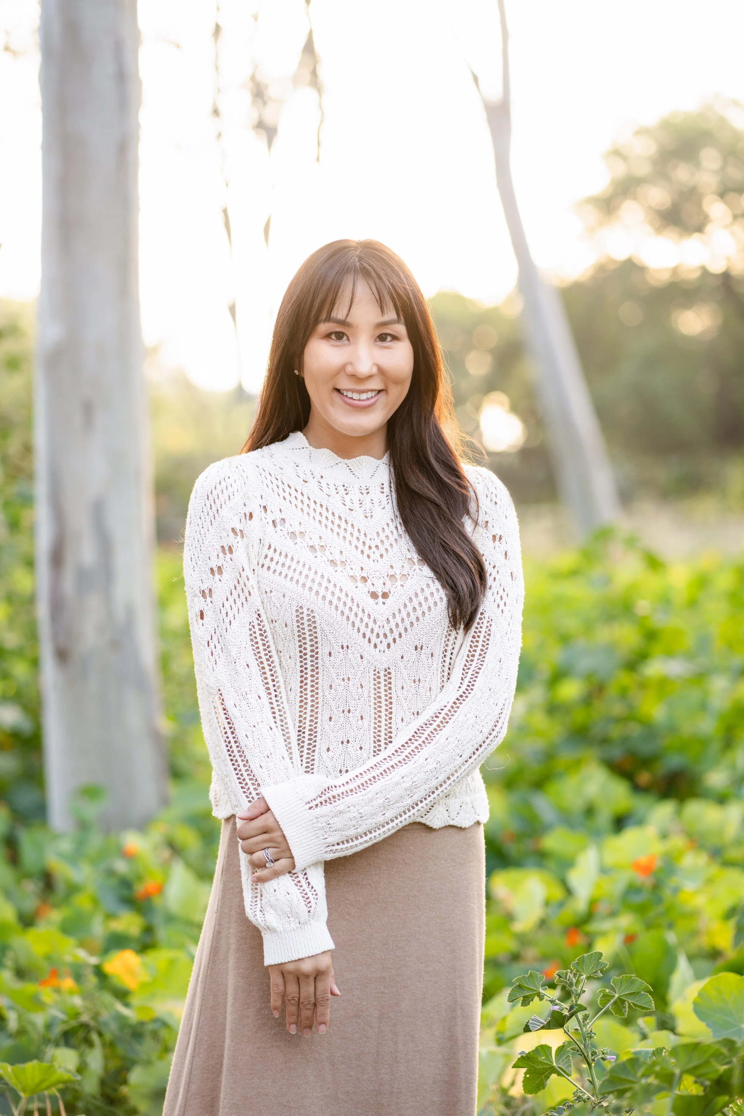 A woman with long dark hair smiling outdoors in a field with tall trees and greenery, wearing a white knitted sweater and a beige skirt during sunset.