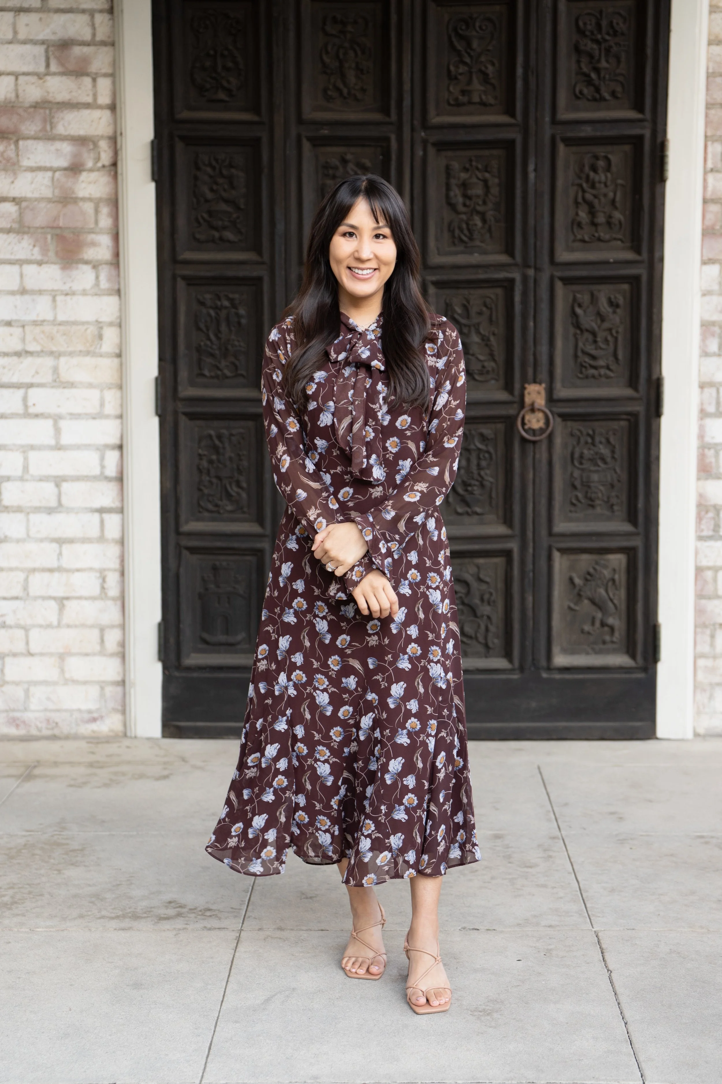 A smiling woman in a long, floral print dress and nude heels stands in front of a carved, dark wooden door with a lion-shaped door knocker, on a brick and concrete porch.