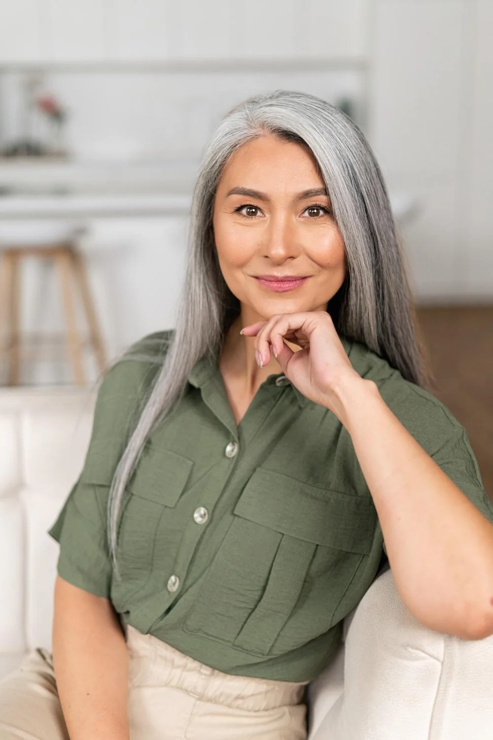 A woman with long gray hair, wearing a green button-up shirt, sitting indoors and smiling at the camera.