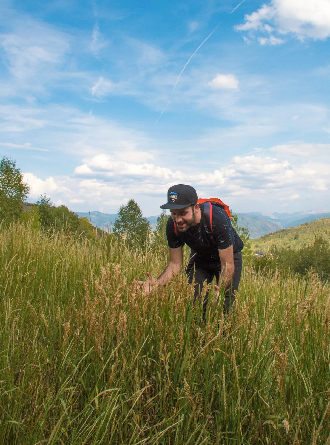 A man with a beard wearing a black cap, black t-shirt, and a red backpack is crouched down in tall grass, smiling as he examines the plants amid a mountain landscape with a partly cloudy sky.