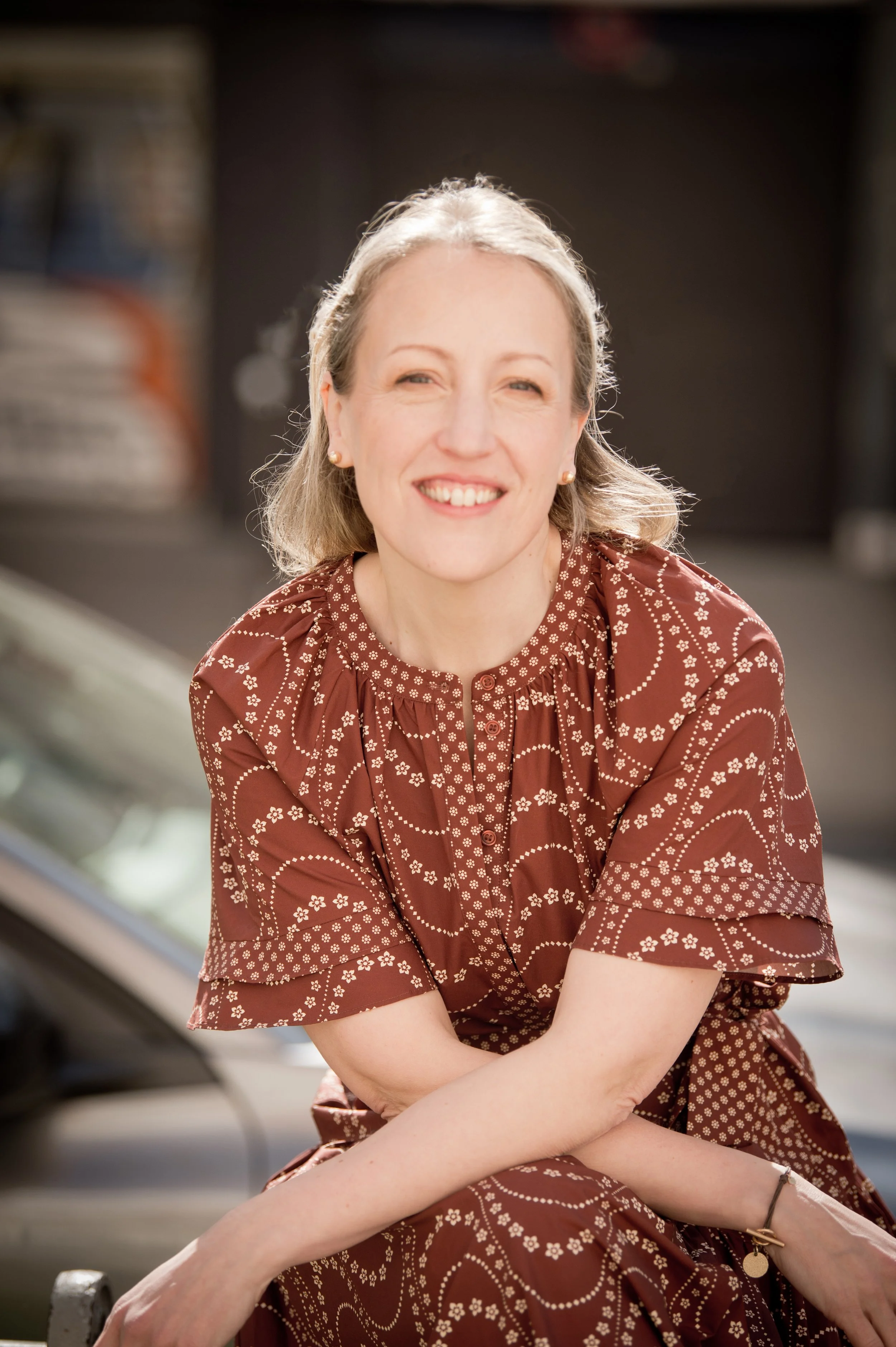 Une femme souriante portant une robe marron avec un motif floral blanc, assise devant une voiture.