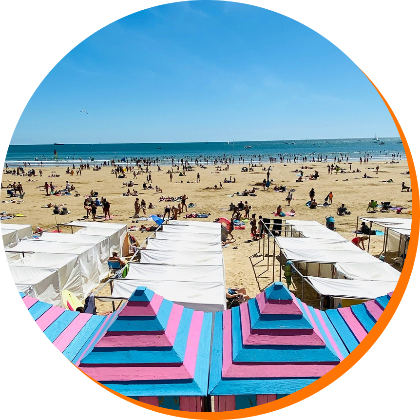 Plage ensoleillée avec de nombreuses personnes, cabanes et parasols, mer bleue avec des bateaux en arrière-plan.