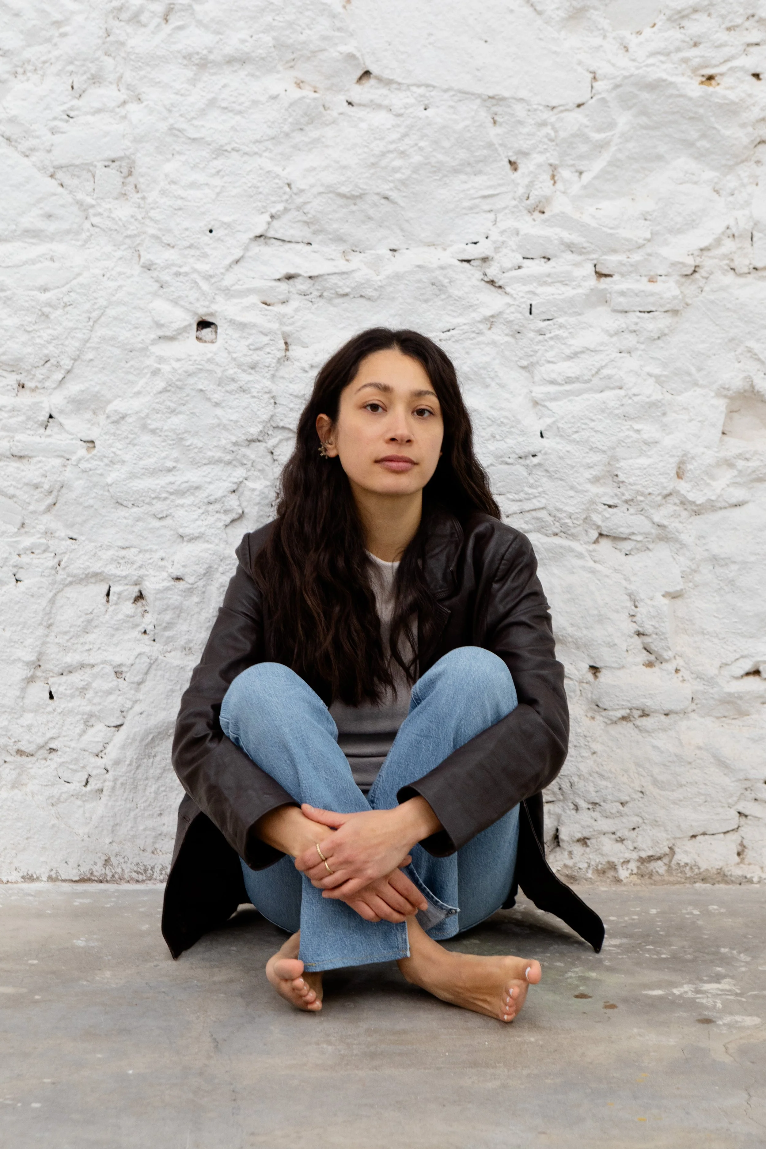 Junge Frau sitzt barfuß vor einer weißen, unregelmäßig gemauerten Wand, trägt Jeans und einen schwarzen Lederjacke.