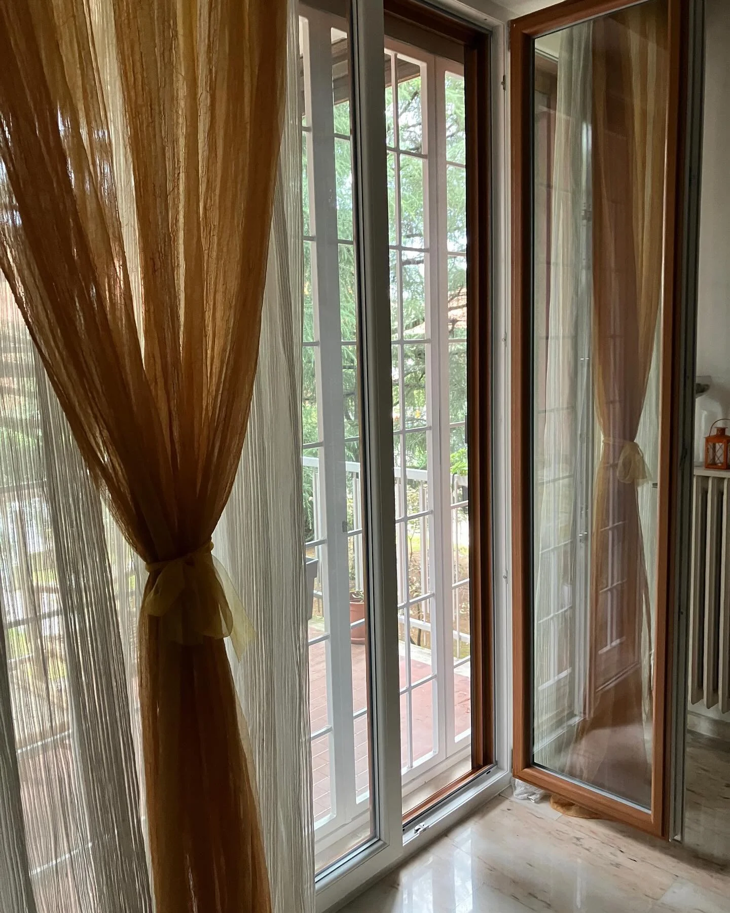 Vista di una porta finestra in legno e vetro con tende arancioni e bianche, che dà su un balcone con piante e alberi verdi.