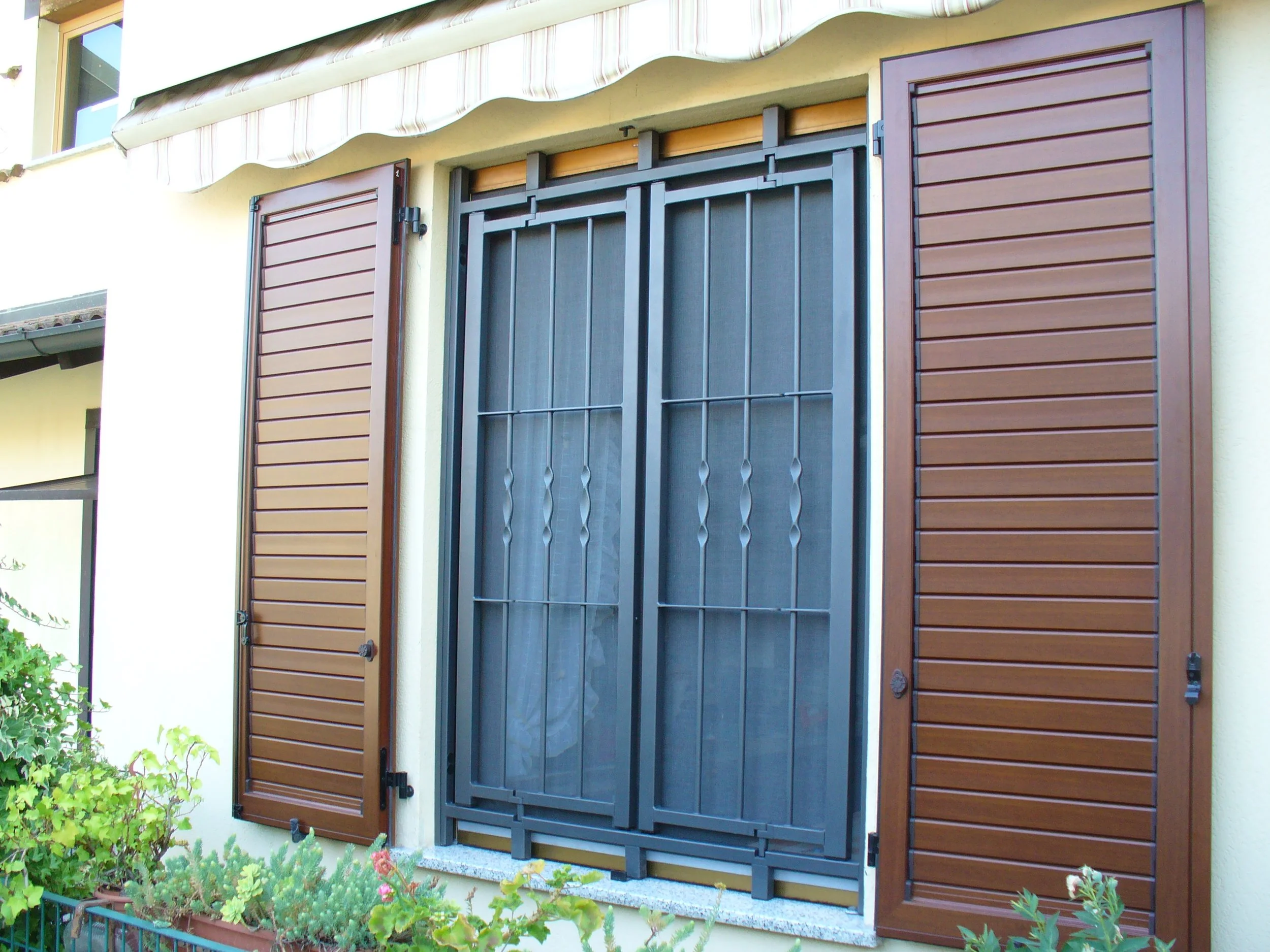 Finestra con persiane in legno e inferriate nere su parete gialla con finestra superiore parzialmente visibile