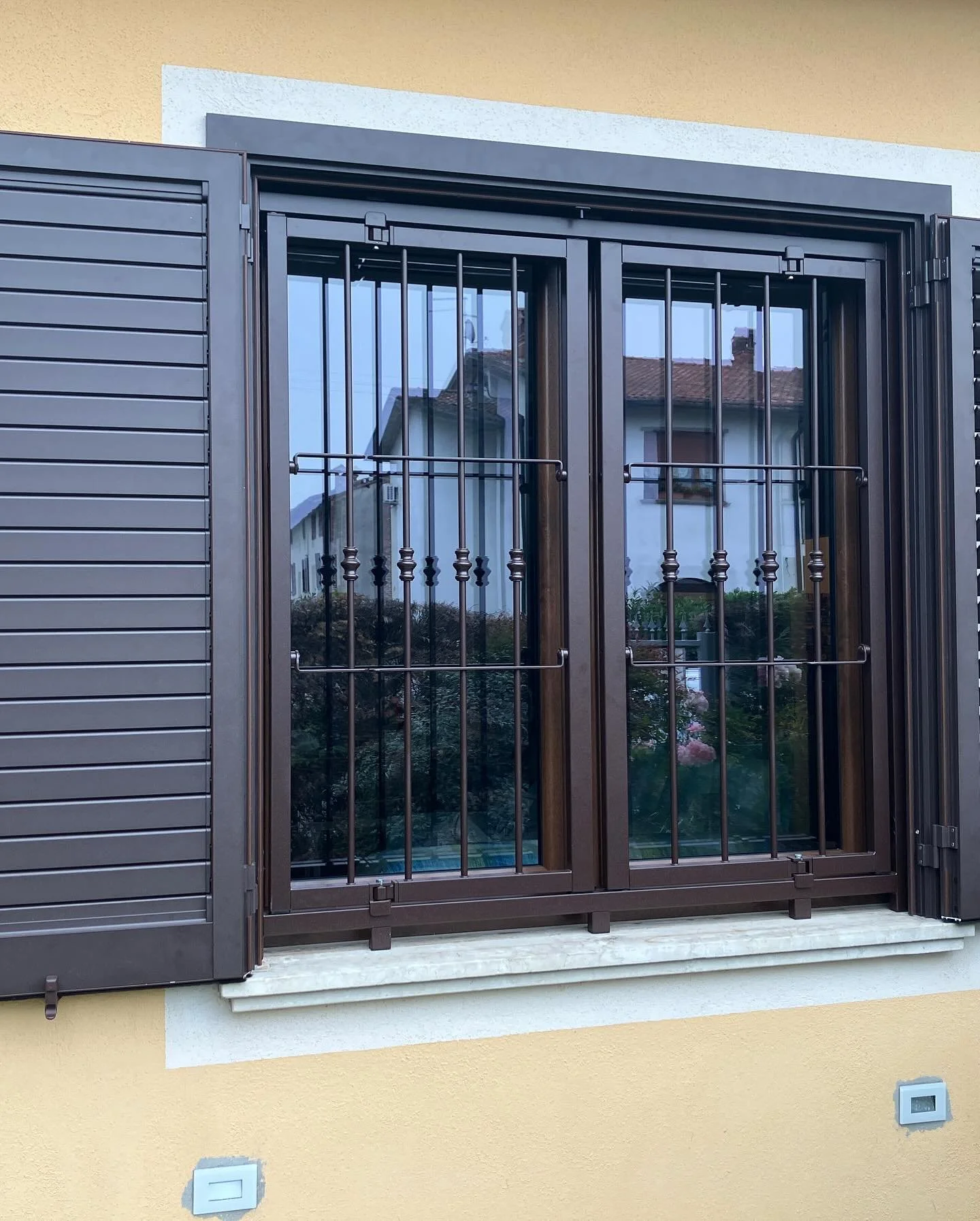 Finestra con inferriate di metallo e persiane di colore marrone sulla facciata di una casa, con riflesso di altre abitazioni e vegetazione.