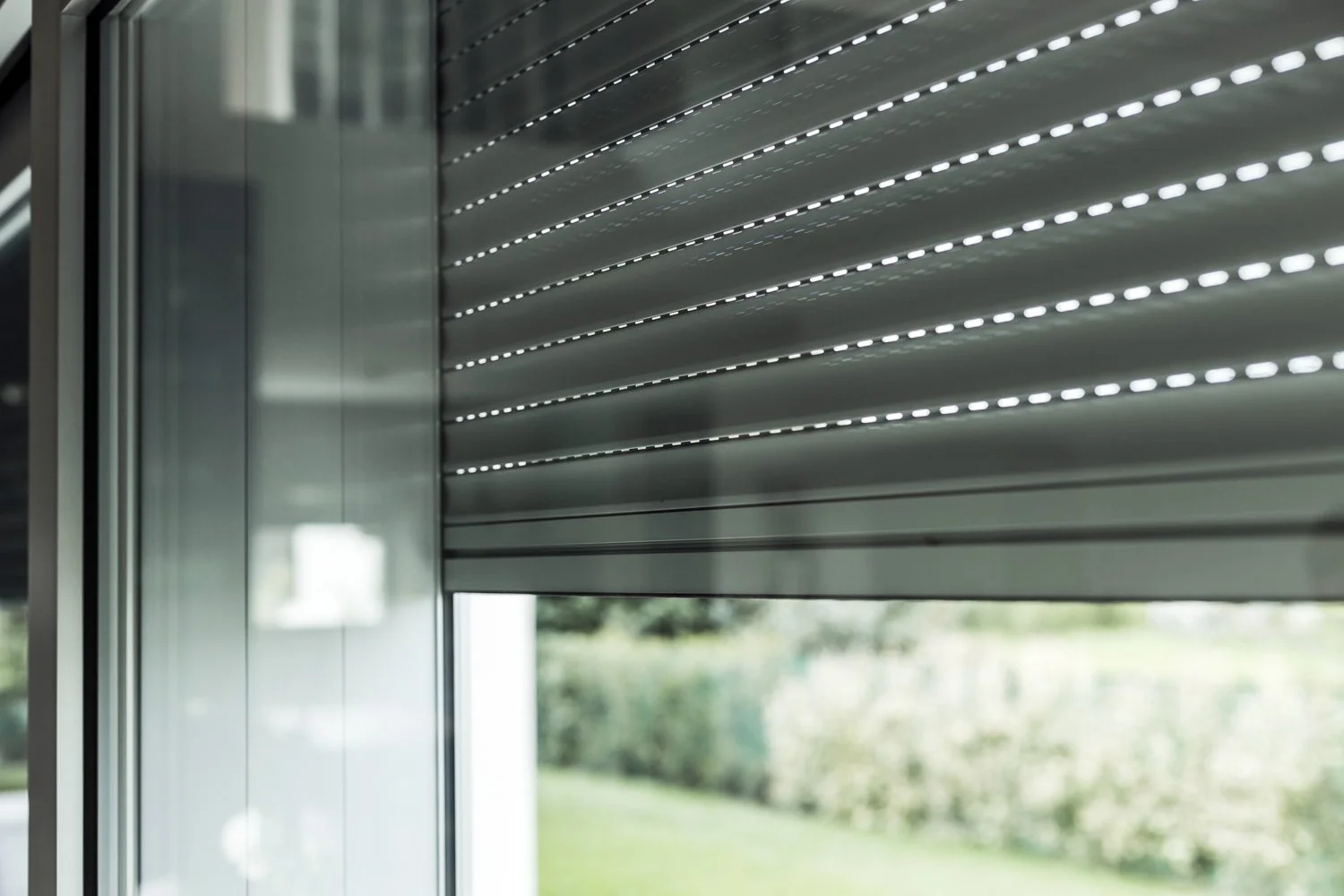 Dettaglio di una finestra con tapparelle di metallo chiuse, con uno scorcio di natura visibile attraverso la parte inferiore dello sfondo.