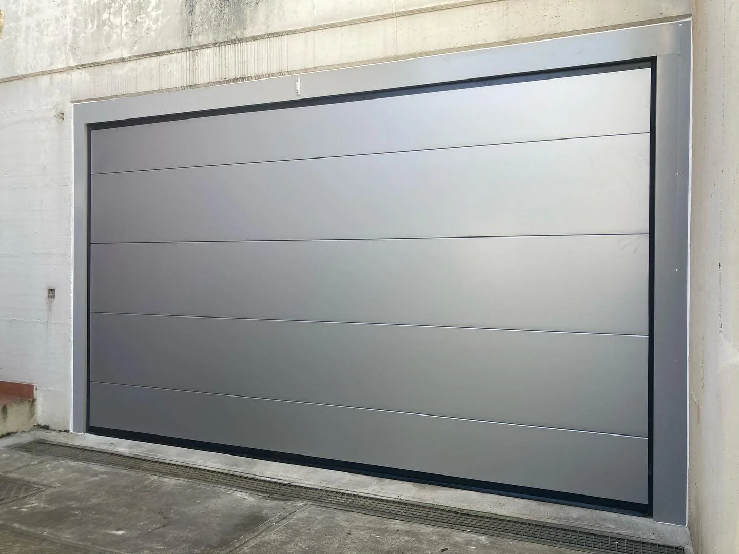 Porta del garage grigia in metallo con pannelli orizzontali, installata tra pareti di cemento, con sistema di drenaggio sulla pavimentazione sottostante.