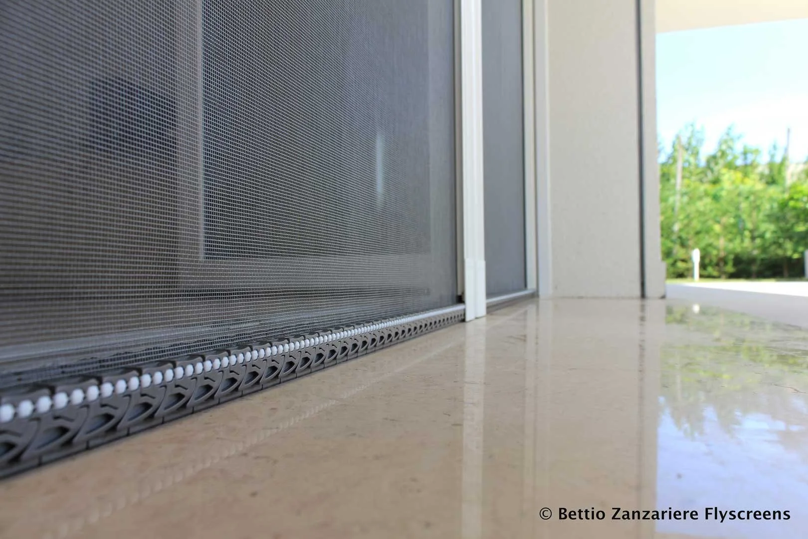 Dettaglio di uno schermo a moschettone installato sulla soglia di una porta o finestra, con sfondo di ambiente esterno con alberi verdi e cielo azzurro.