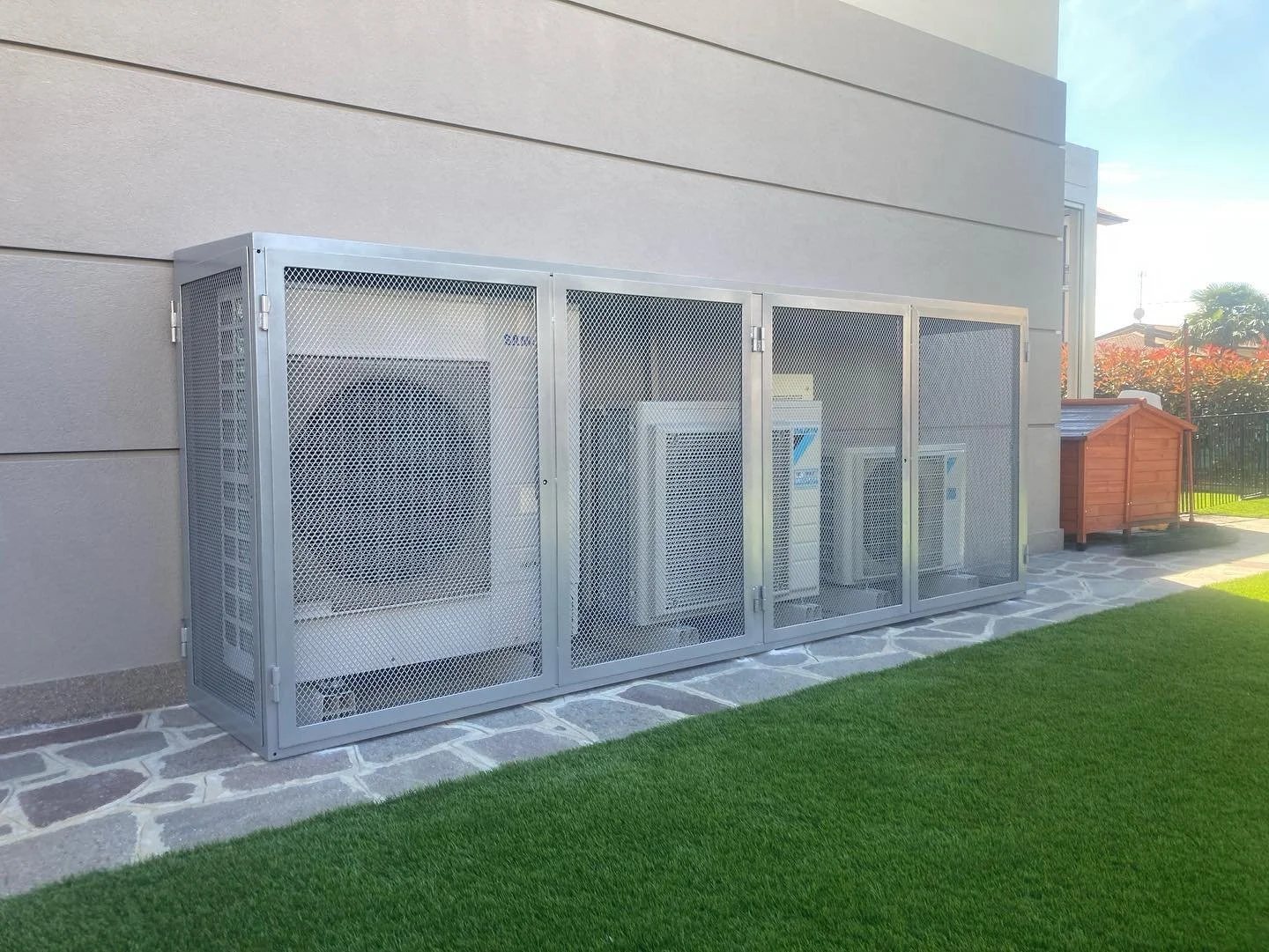 Armadio di metallo con grille di protezione, collocato sul lato di una casa, con un prato verde e una piccola casetta di legno sullo sfondo.