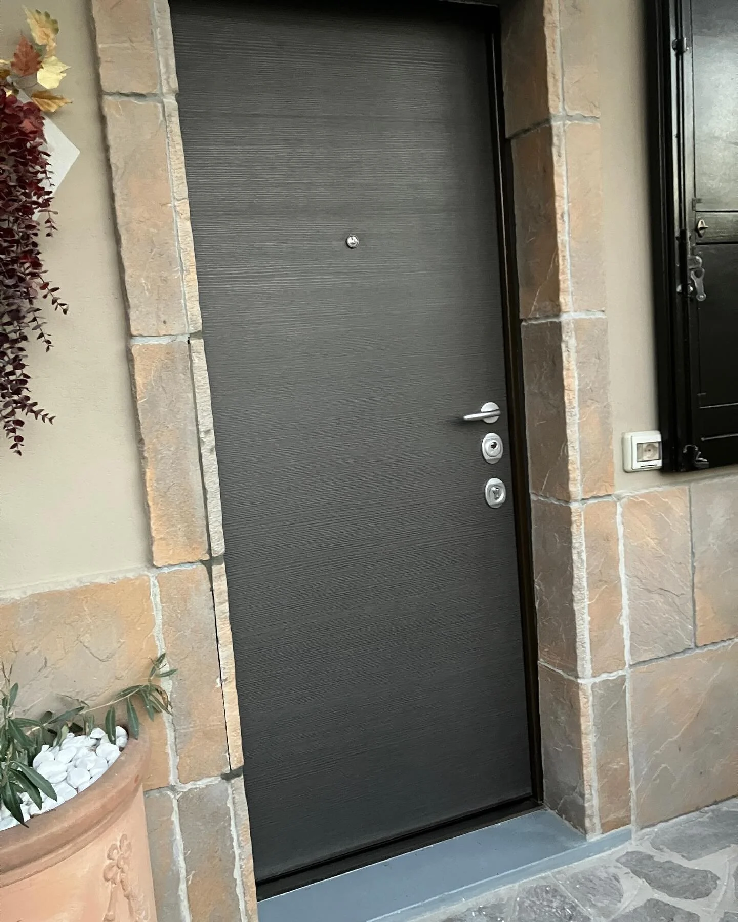 Porta d'ingresso nera con maniglia e cilindri di sicurezza, circondata da pietre beige e decorazioni verdi