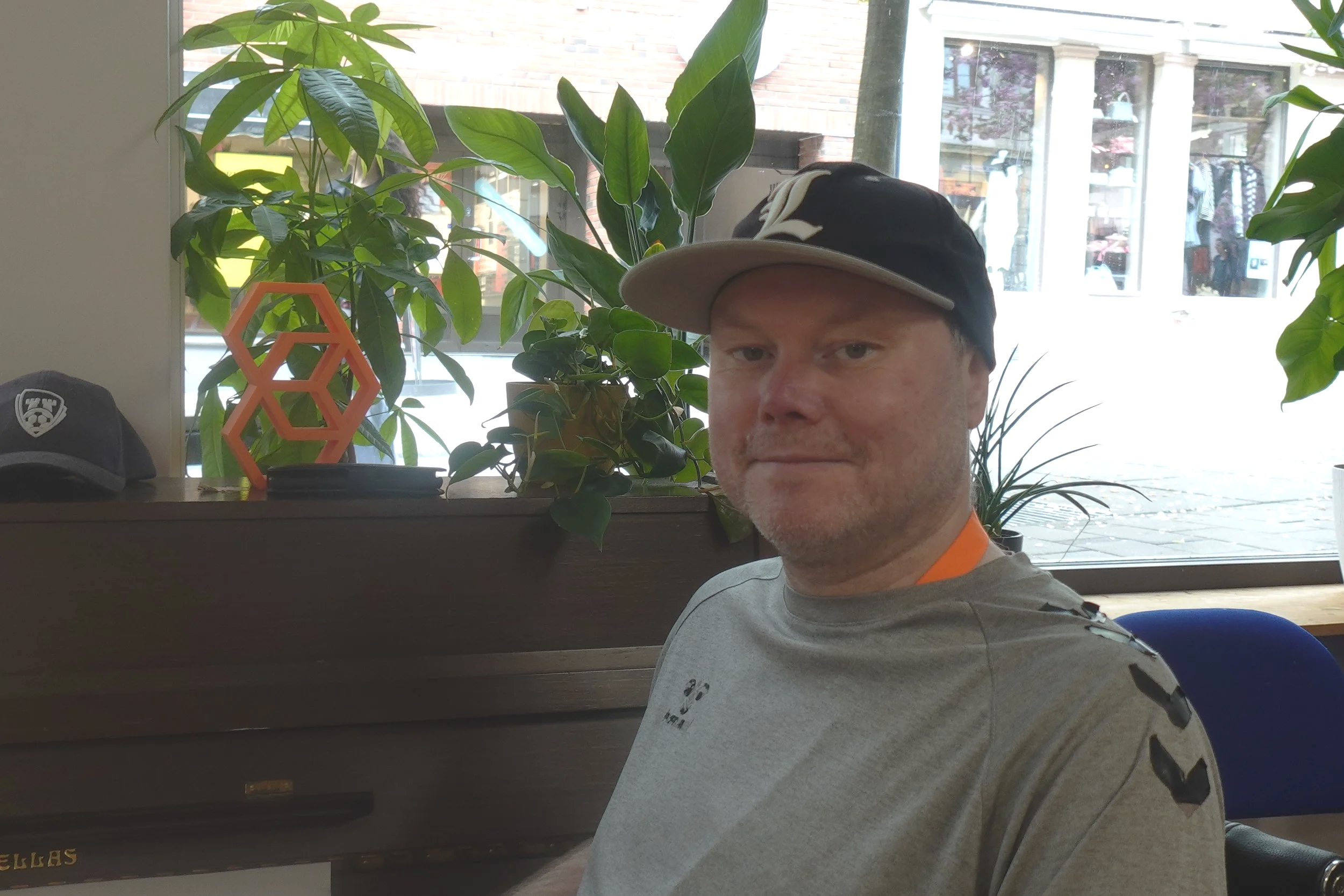 Et menneske tar et selfiesyn med en bakgrunn av grønne planter og en stor vindusglass som viser en gate med butikker og mennesker utenfor.